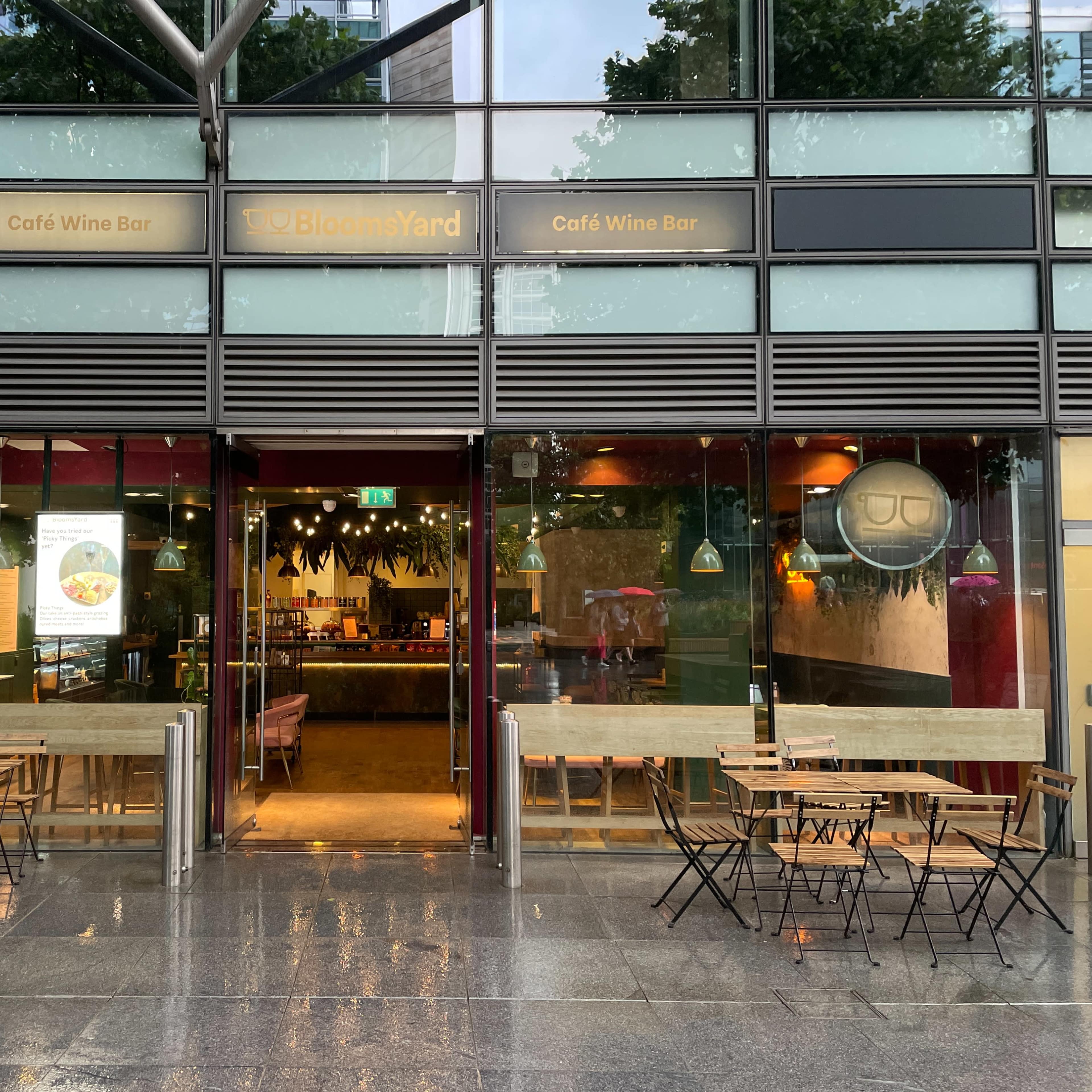 The exterior of a café and wine bar with glass doors and tables set outside, located in a modern building during a rainy day.