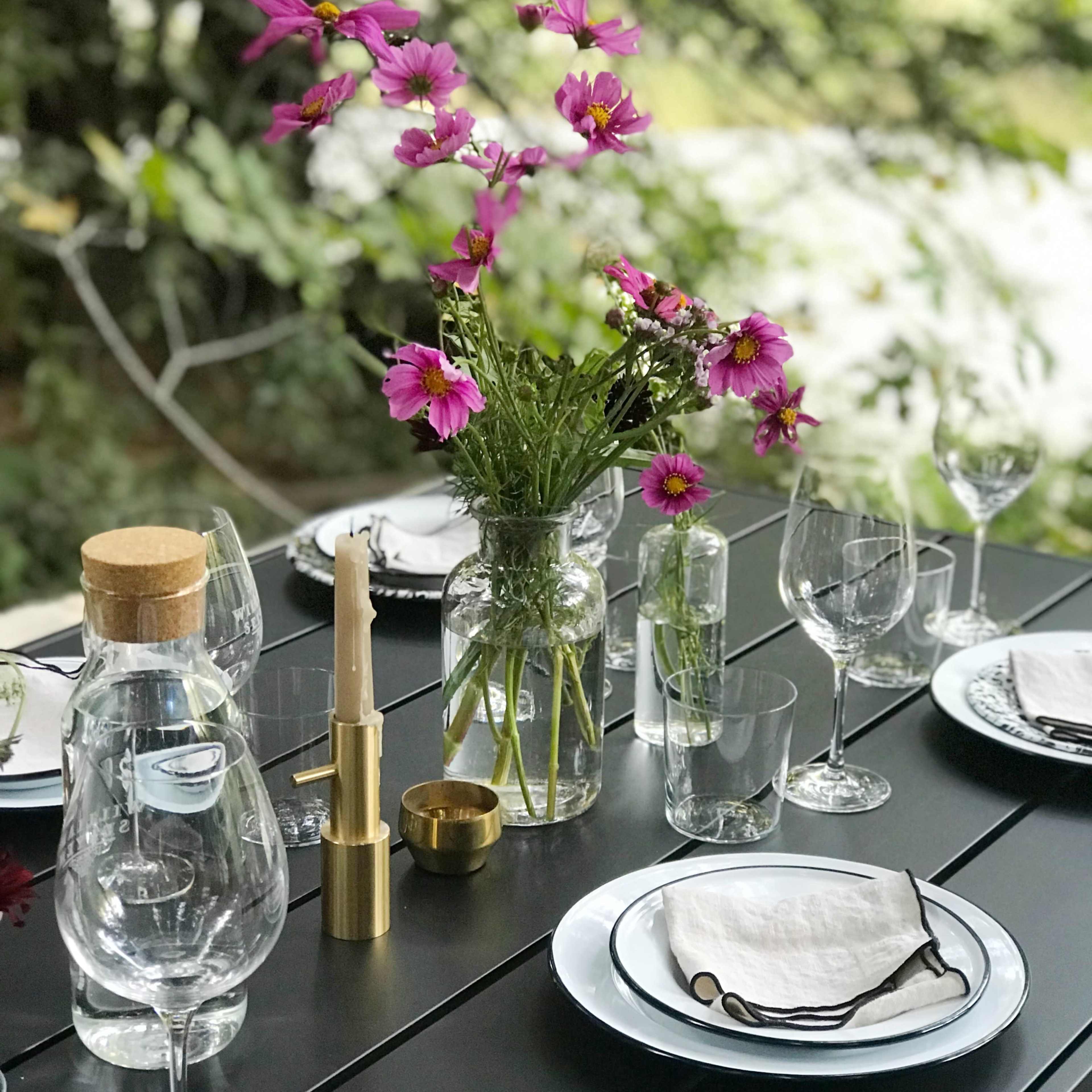 A dining table is set outdoors with clear glassware, white plates, and a vase of pink flowers as a centerpiece.