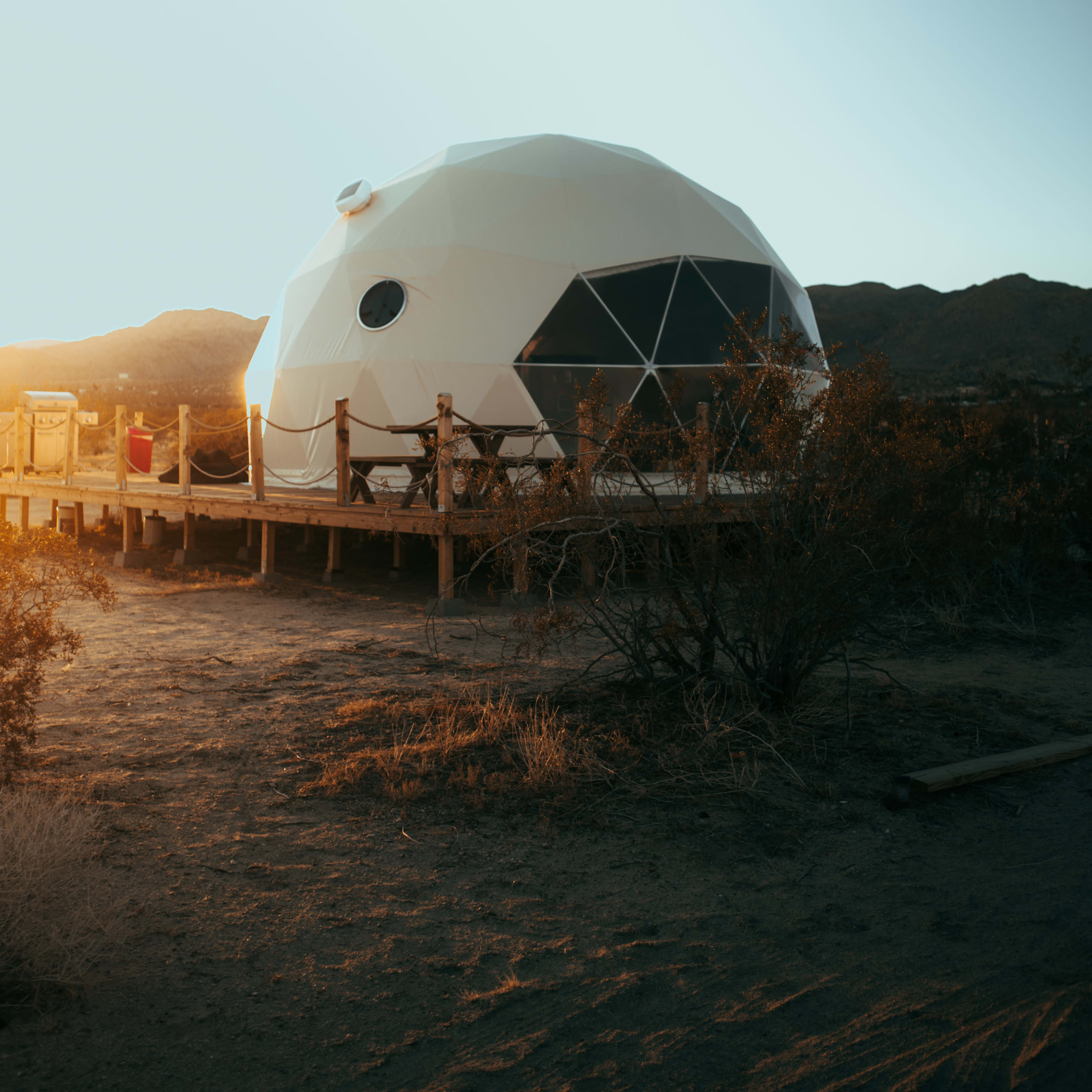 A geodesic dome structure is situated on a wooden deck in a desert landscape with mountains in the background during sunset.