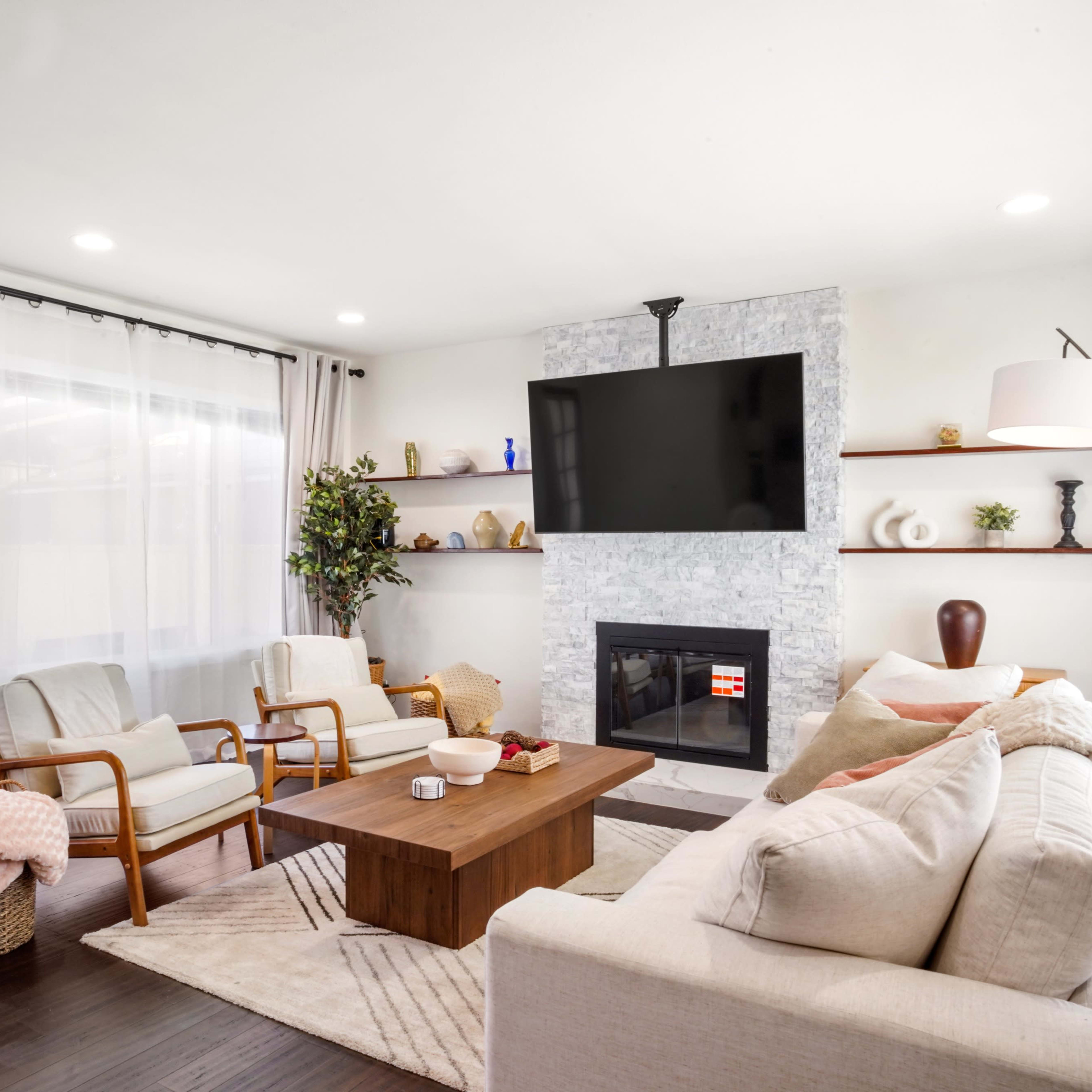 A modern living room with a stone fireplace, a wall-mounted TV, a coffee table, and seating arrangements with two chairs and a sofa.