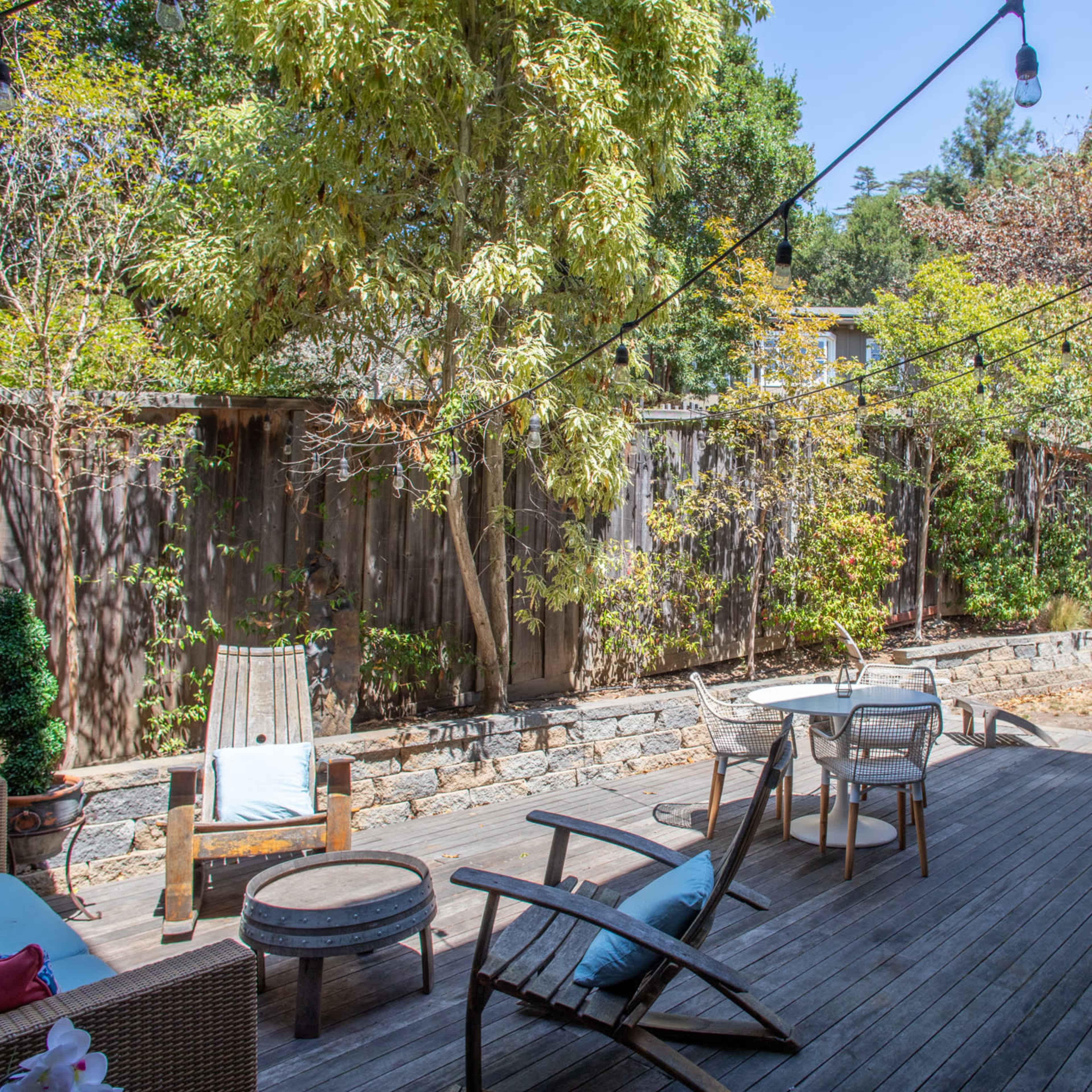 A wooden deck features outdoor seating and a fire pit, surrounded by a landscaped yard with trees and overhead string lights.