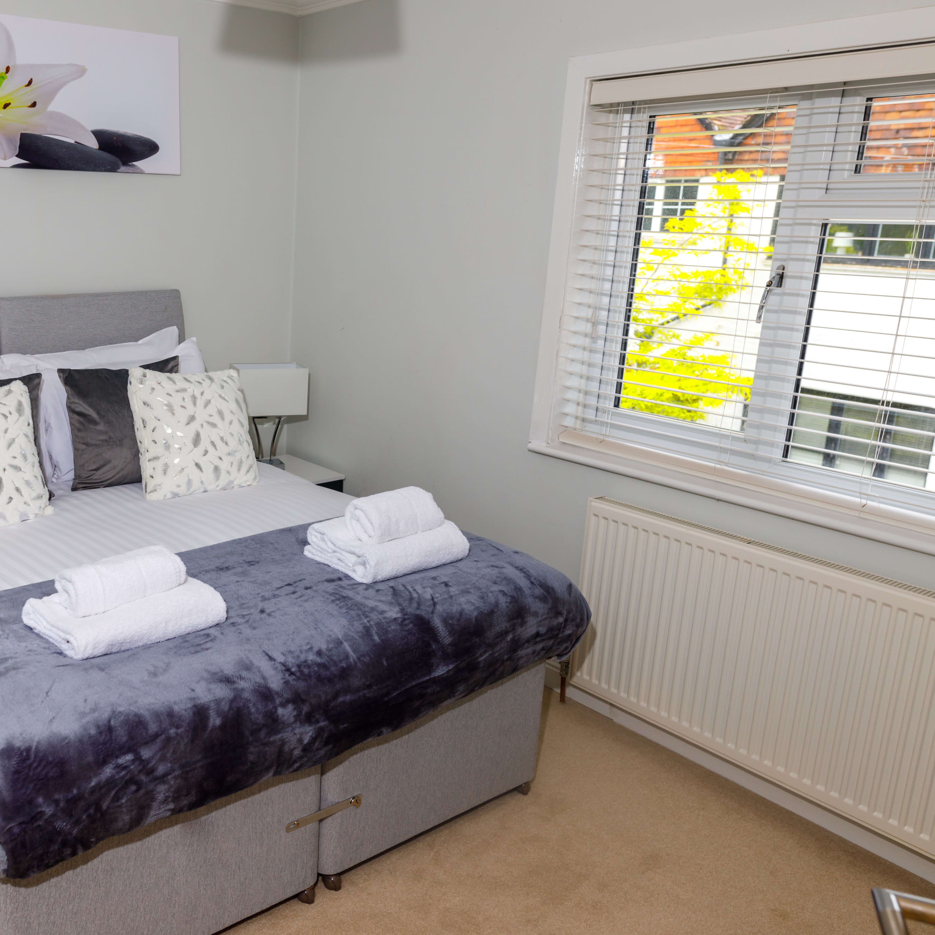 A modern bedroom features a neatly made bed with gray and white linens, decorative pillows, and fresh towels on a plush bedspread.
