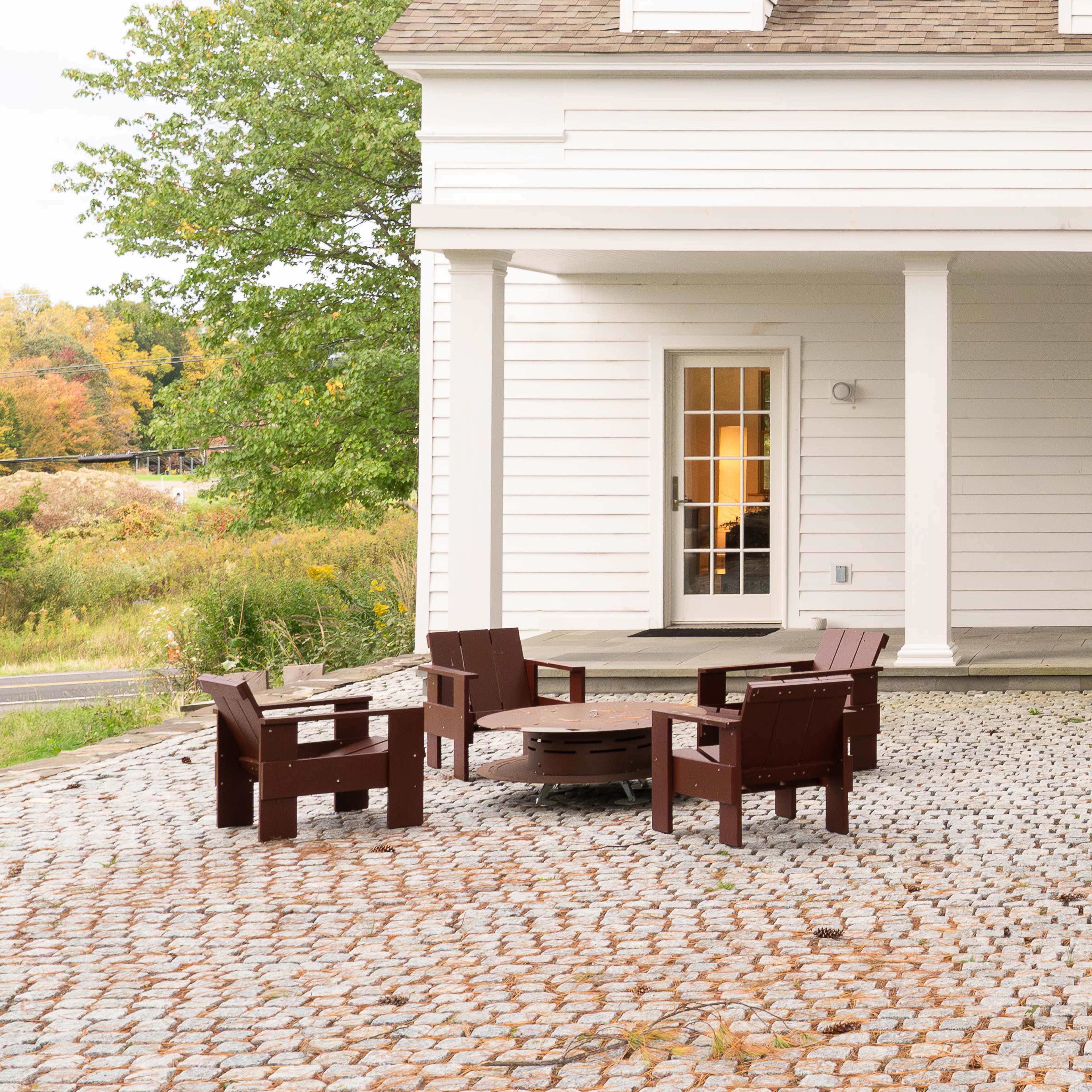 A seating area with brown wooden chairs surrounds a fire pit in front of a white house with a sloped roof.