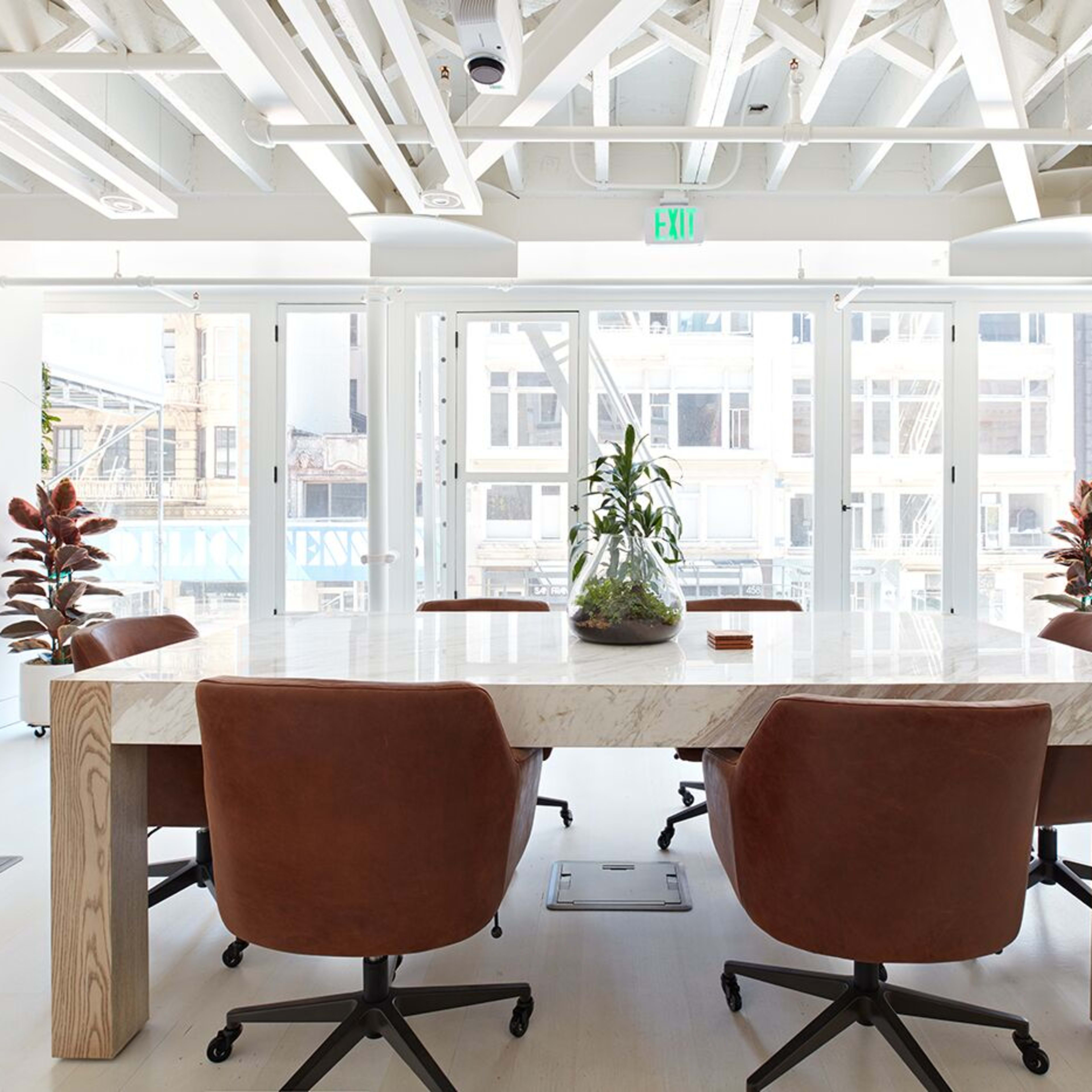 A spacious conference room features a large wooden table surrounded by brown leather chairs, with large windows allowing natural light and views of the city outside.