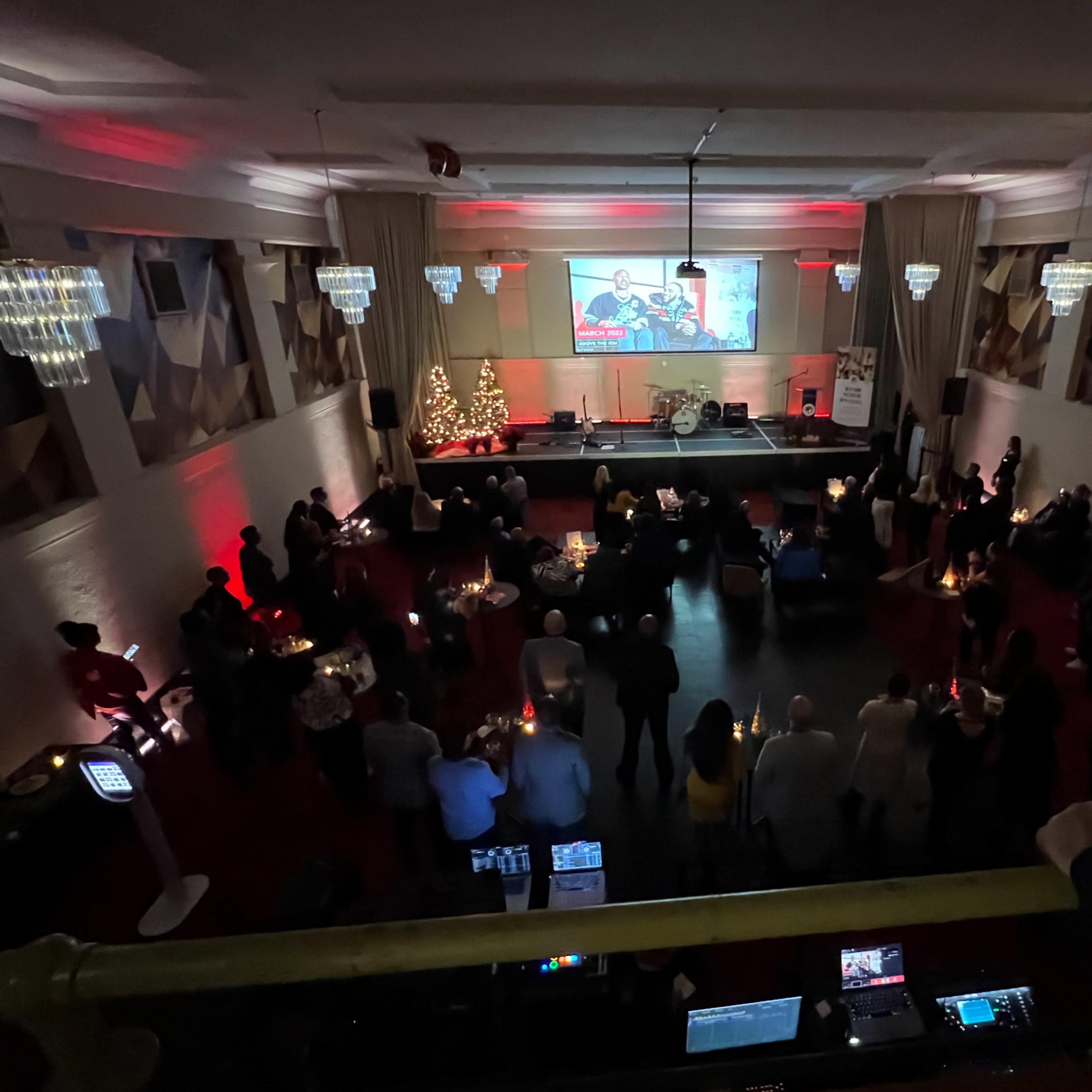 A large group of people gather in a dimly lit venue with a Christmas tree and a screen displaying a festive image.