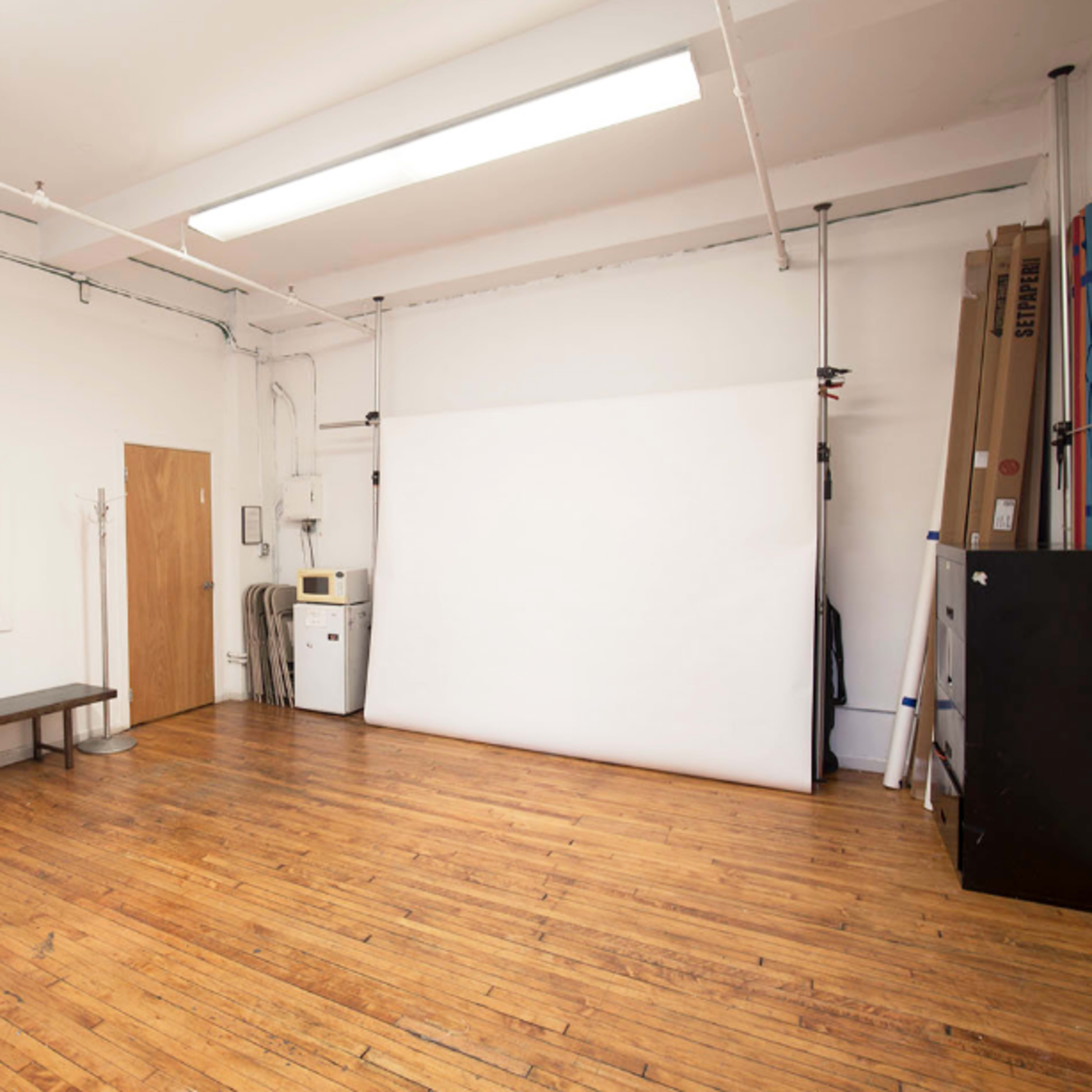 A spacious room with wooden floors, a white backdrop on a wall, and minimal furniture including a bench and a visible doorway.