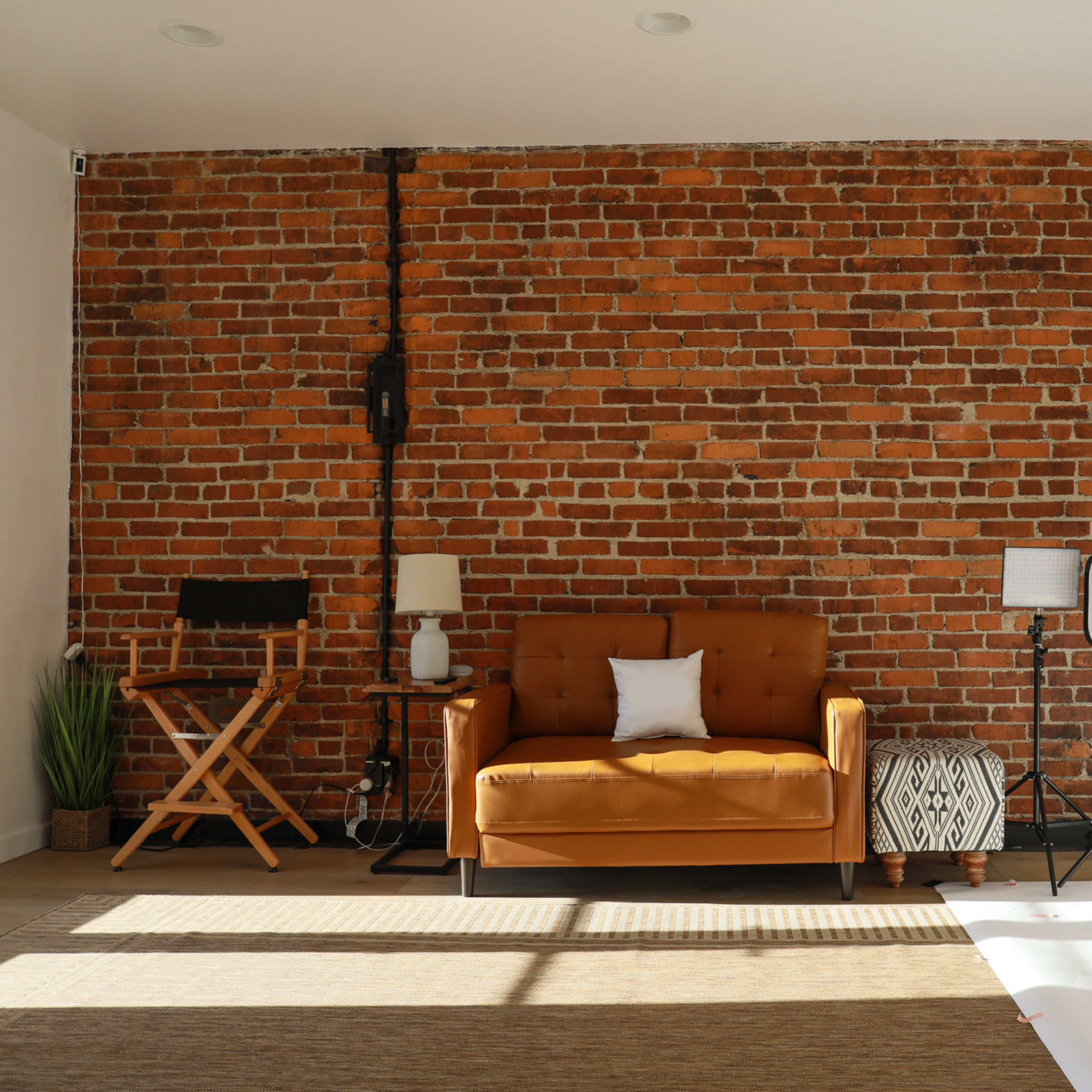 A cozy studio space featuring a brown couch, plants, and studio lights against a brick wall.