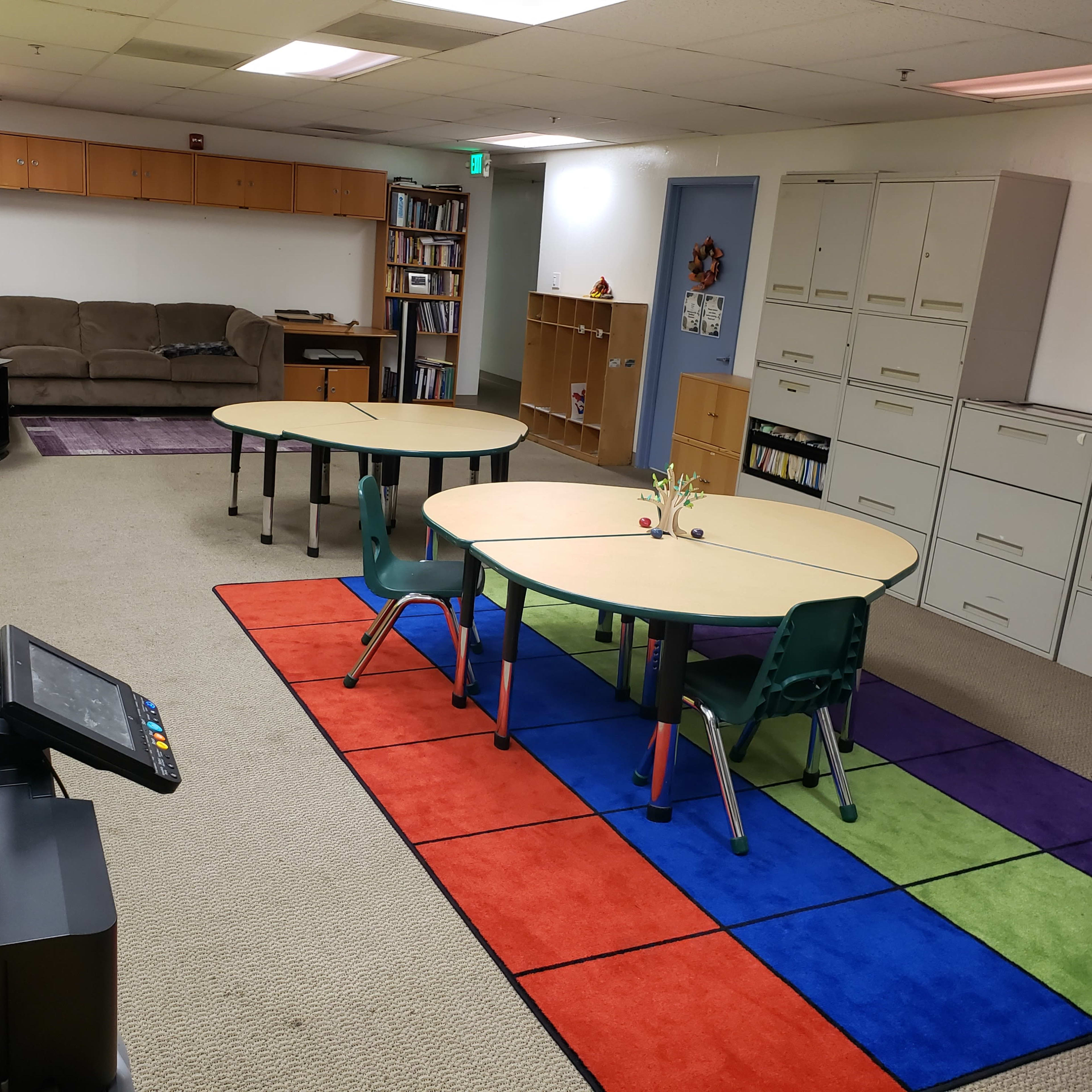A classroom with two circular tables and chairs on a colorful carpet, alongside storage cabinets and a couch in the background.