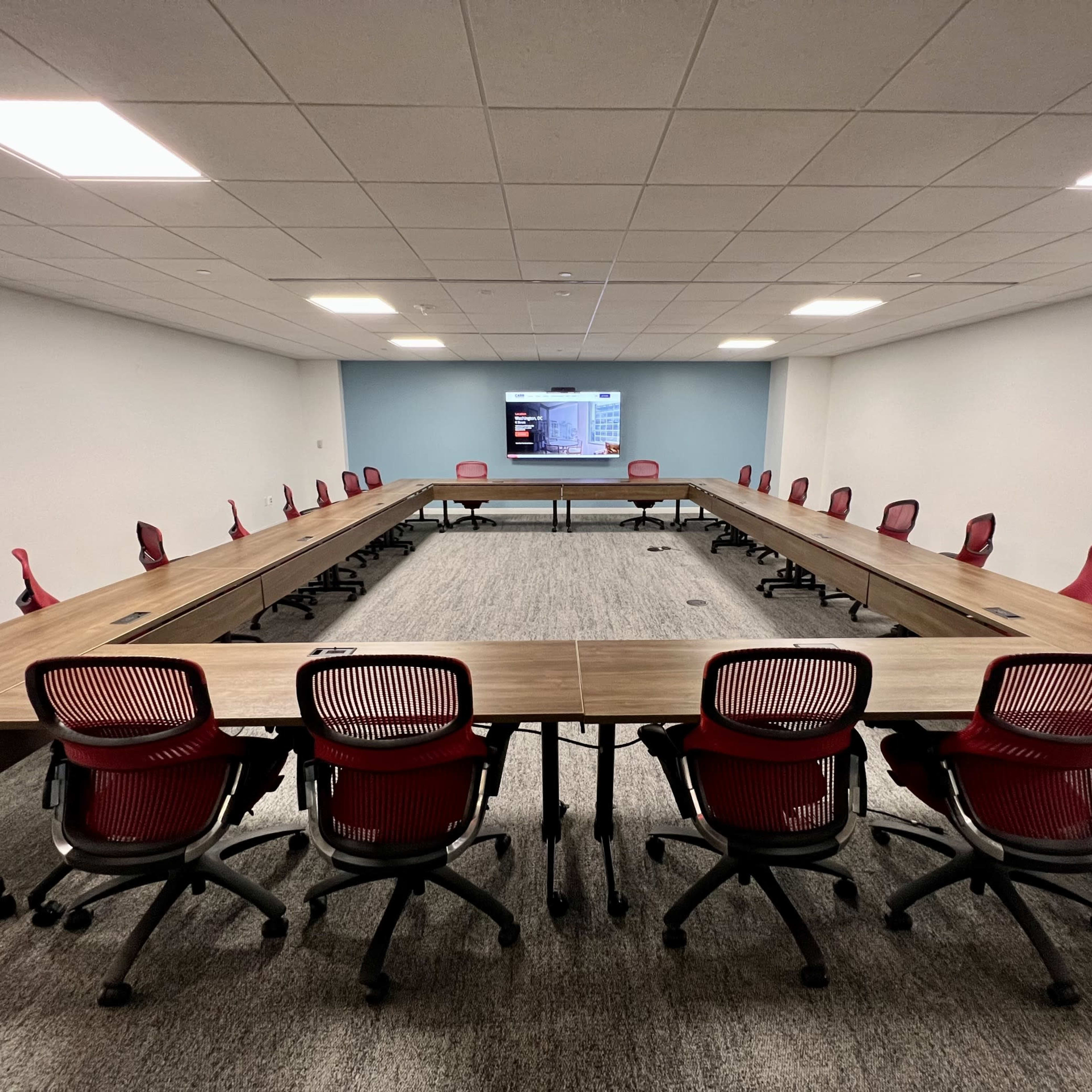 A large conference room features a U-shaped arrangement of wooden tables surrounded by red chairs, with a screen mounted on the wall at one end.