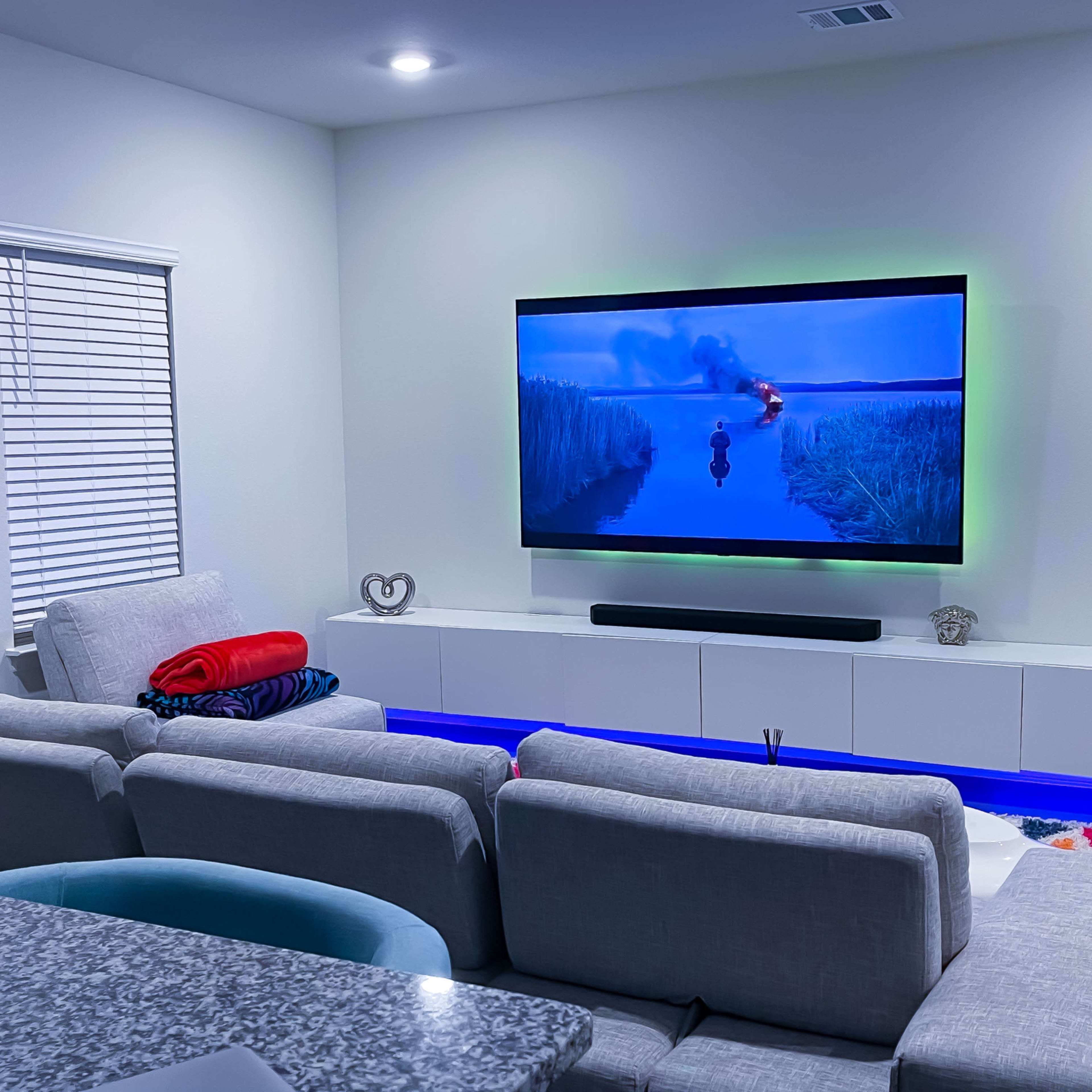 A modern living room features a large television displaying a scene from a movie, with a grey sectional sofa and a lighted entertainment unit underneath.