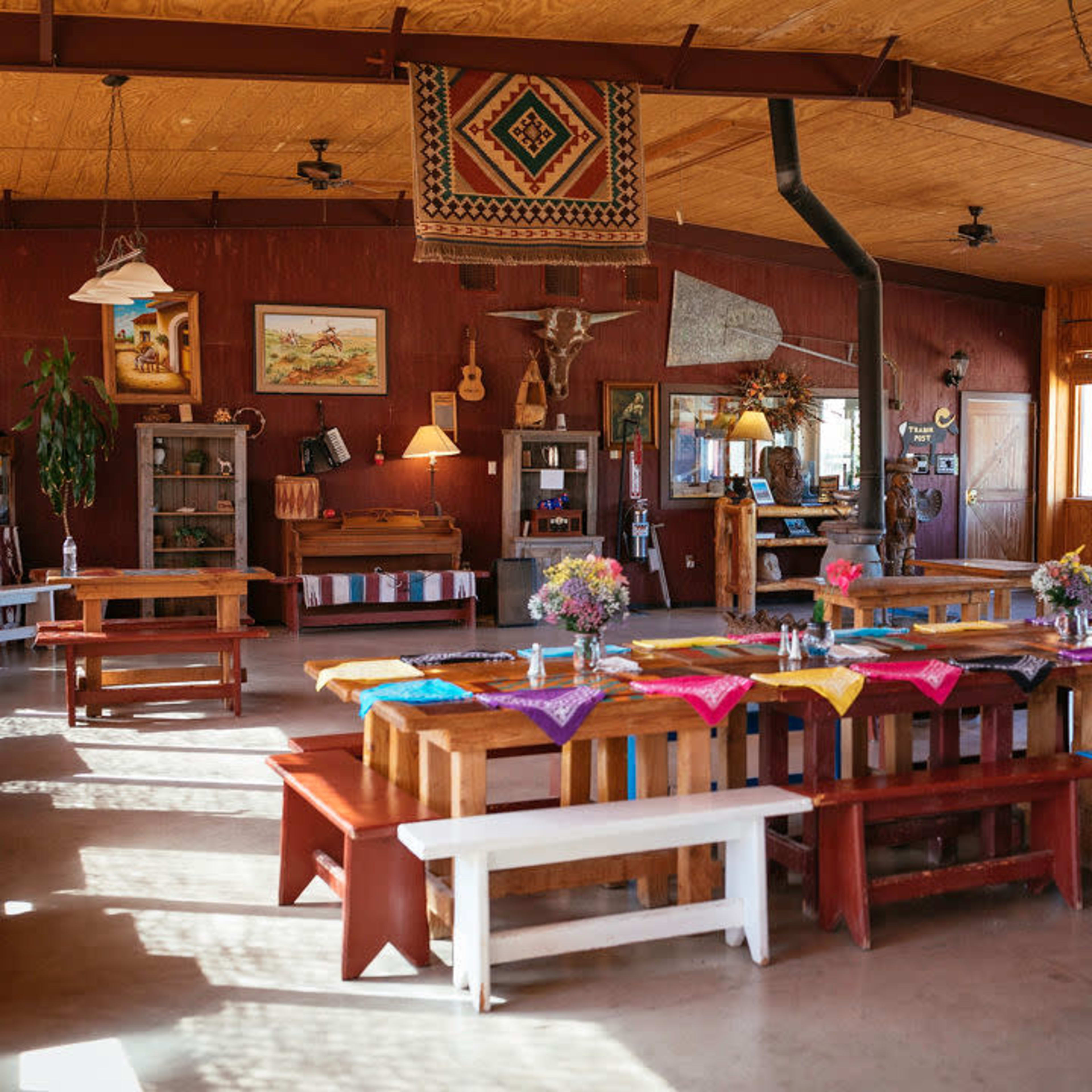 The interior of a rustic dining hall features multiple long wooden tables adorned with colorful table runners and centerpieces, illuminated by natural light from large windows.