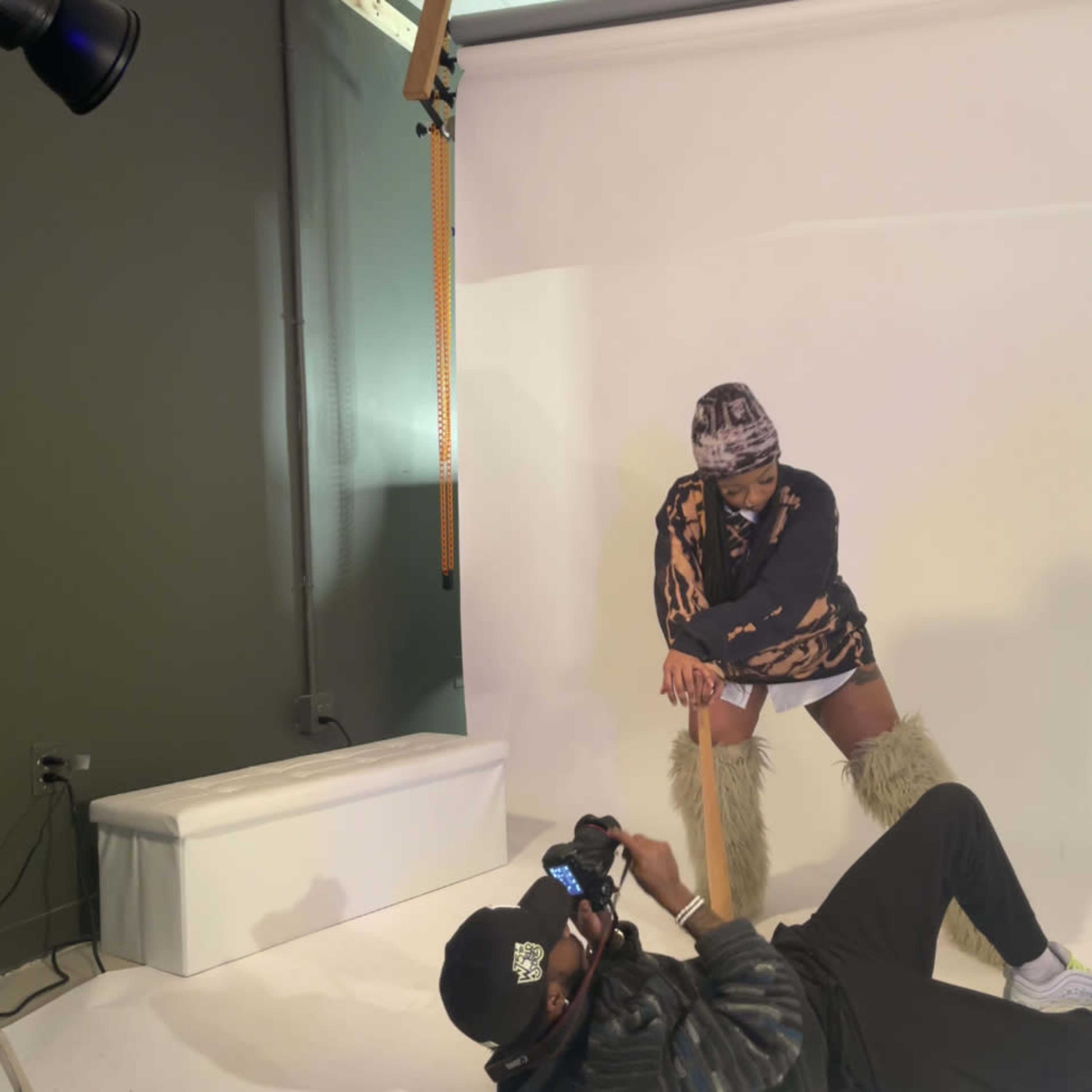 A person poses on a white backdrop while another person lies on the ground taking photos.