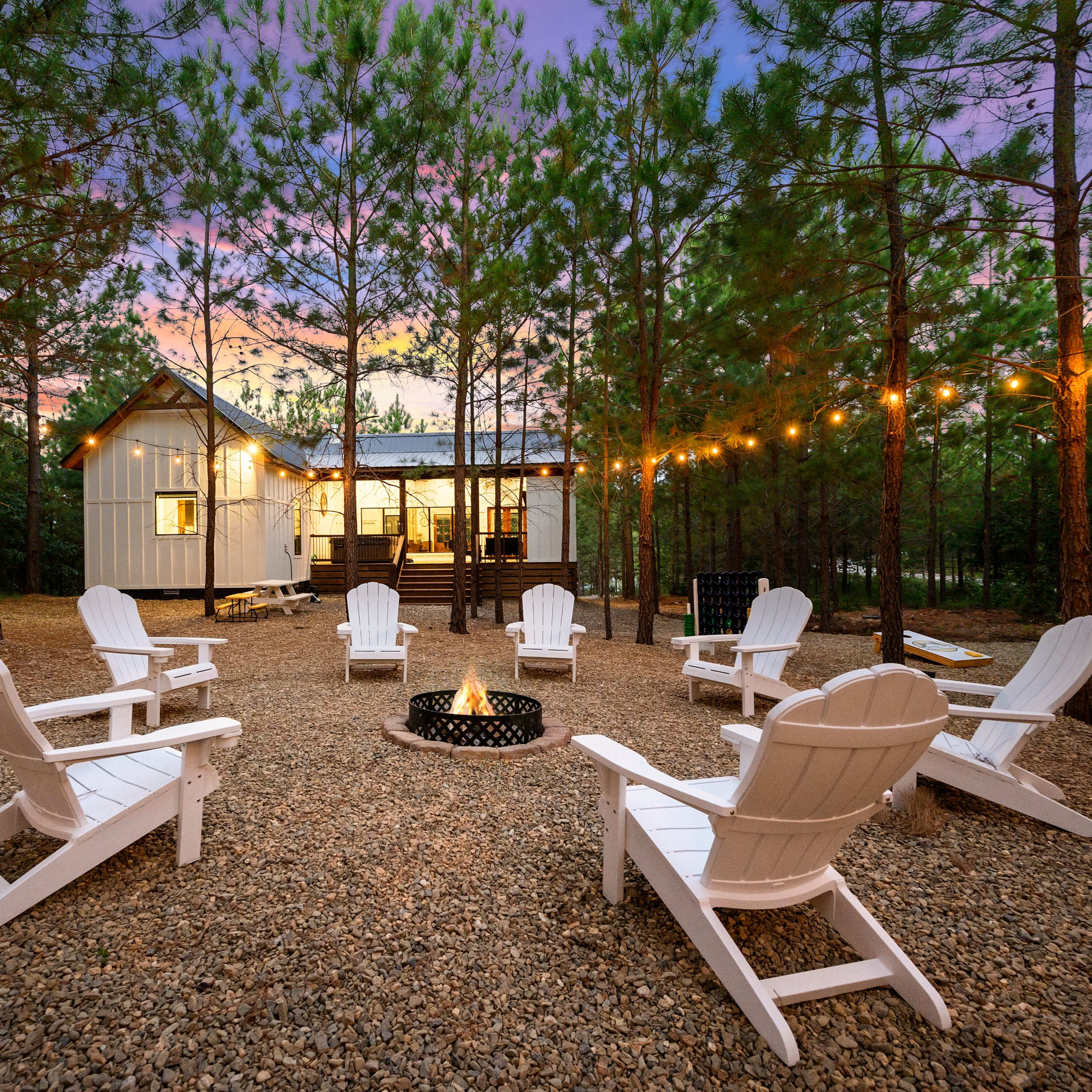 A gravel area surrounded by trees, featuring a fire pit and several white Adirondack chairs arranged in a circle, with a cabin illuminated in the background.