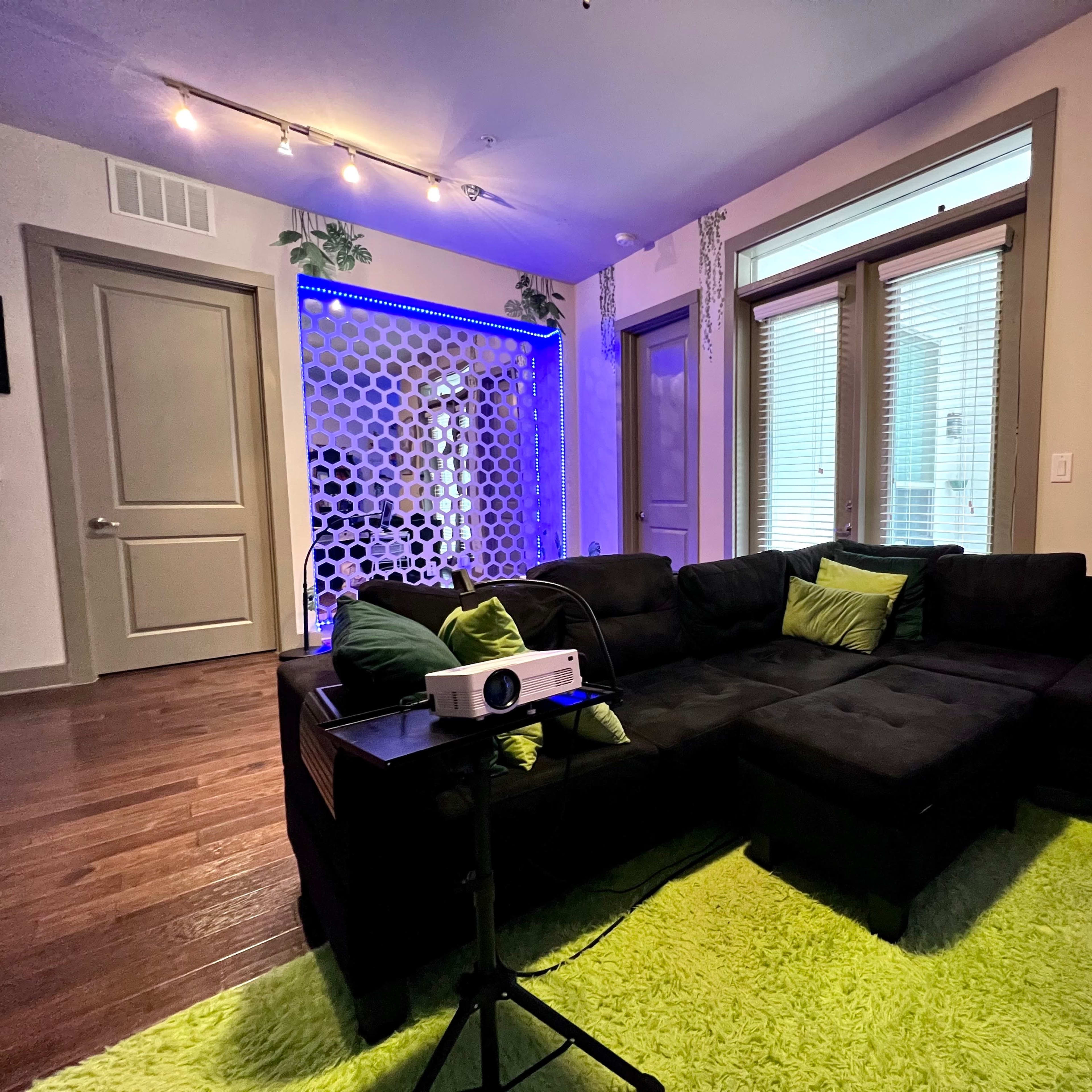 A living room with a black sectional sofa, a green area rug, and a projector on a small table, set against a backdrop of blue-lit walls and a decorative screen.