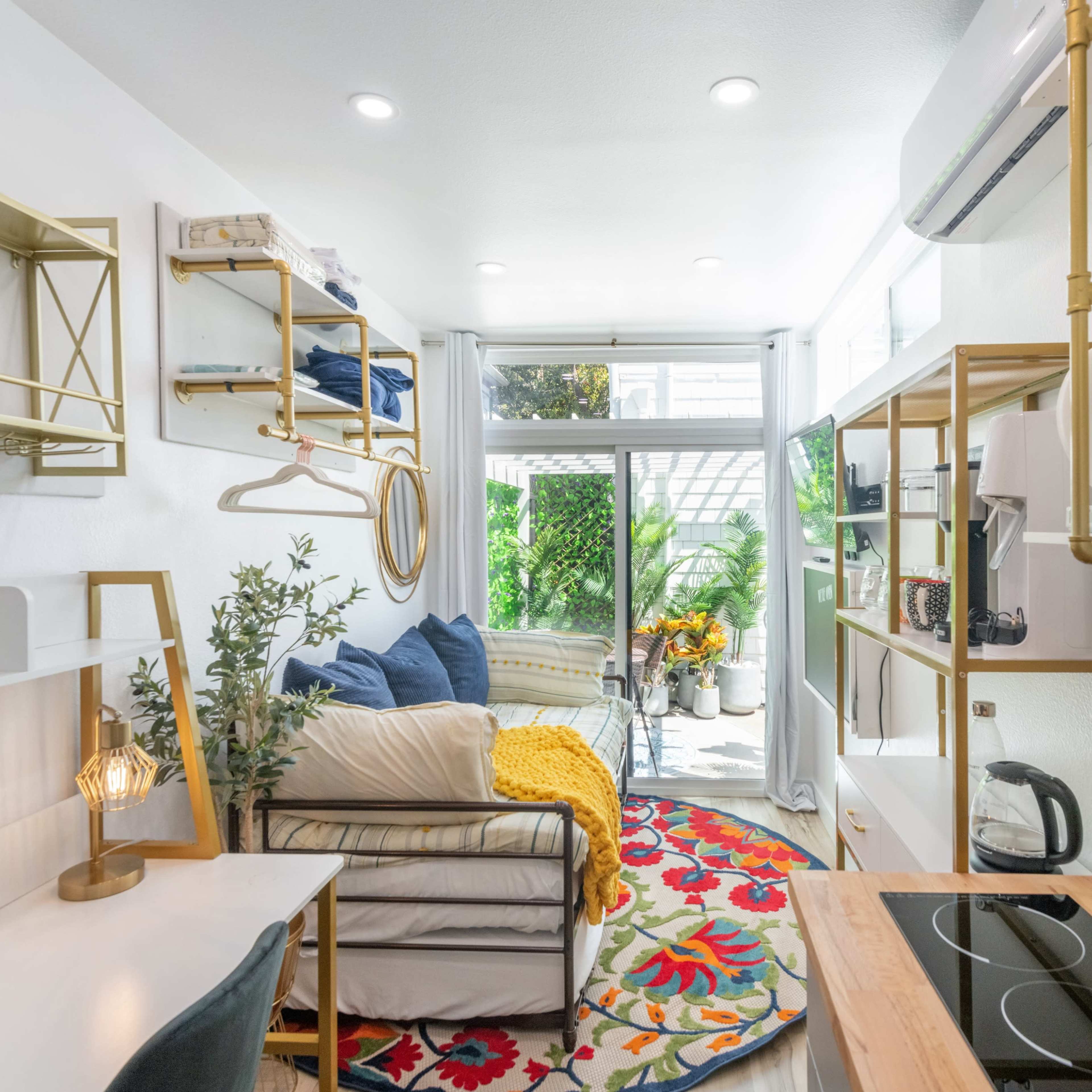 A compact living space featuring a small kitchen area, a workspace with a desk, a seating area, and large windows that open to a patio filled with greenery.