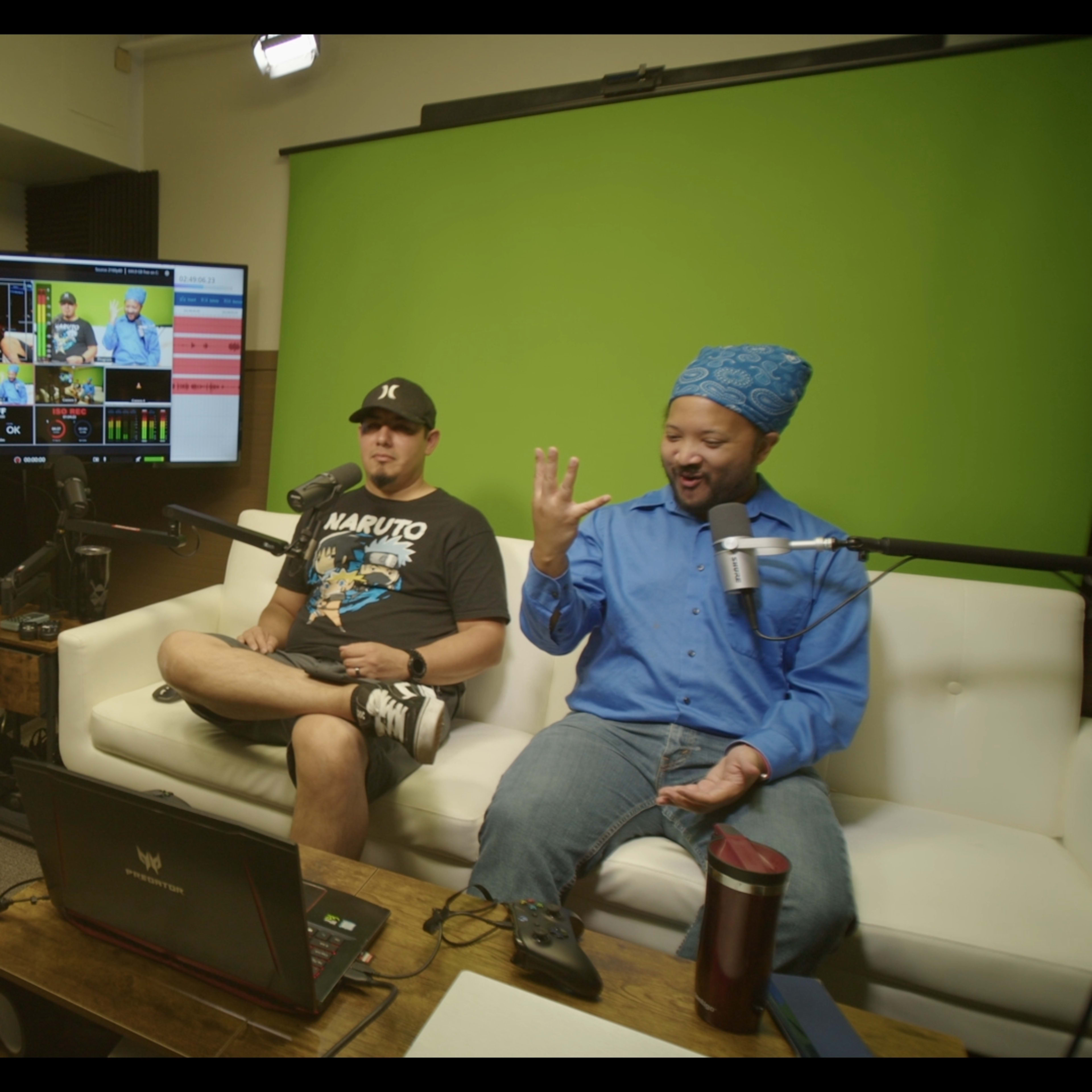Two men are sitting on a white couch in a studio setting, with a green screen backdrop and multiple screens displaying video content in front of them.