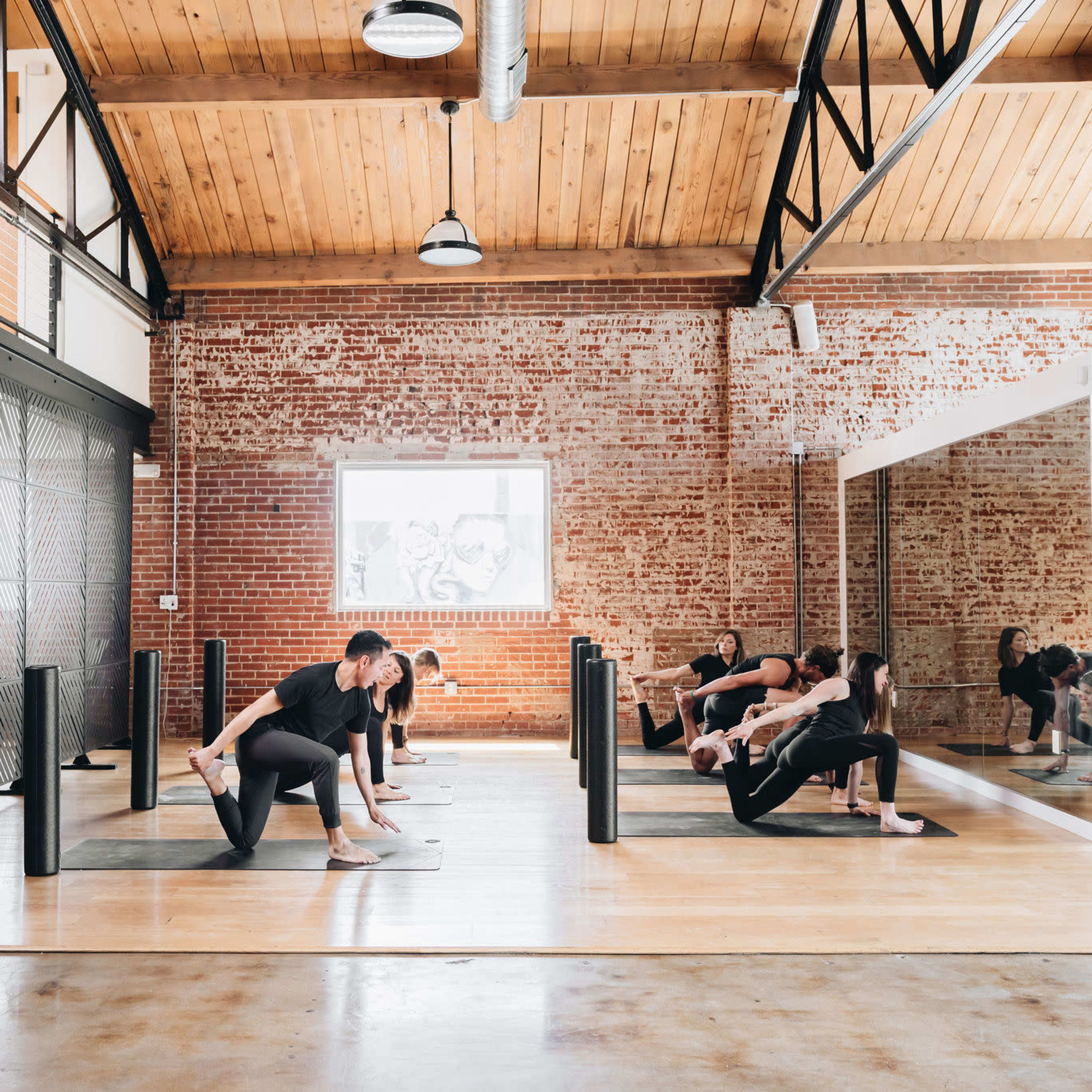 A group of people are practicing yoga in a spacious studio with exposed brick walls and large mirrors.