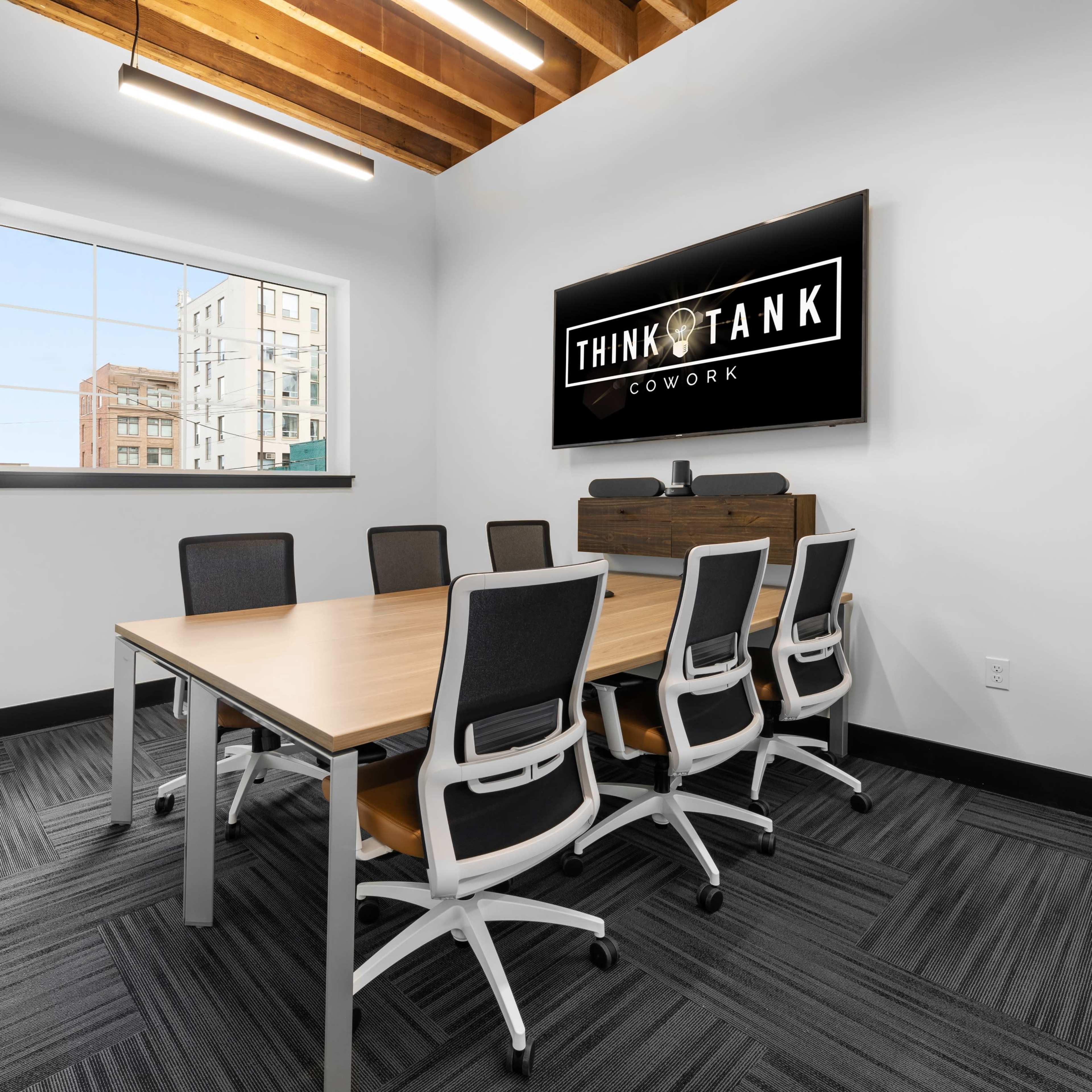 A modern conference room features a long wooden table surrounded by white chairs, with a large screen displaying the "THINK TANK COWORK" logo on the wall.