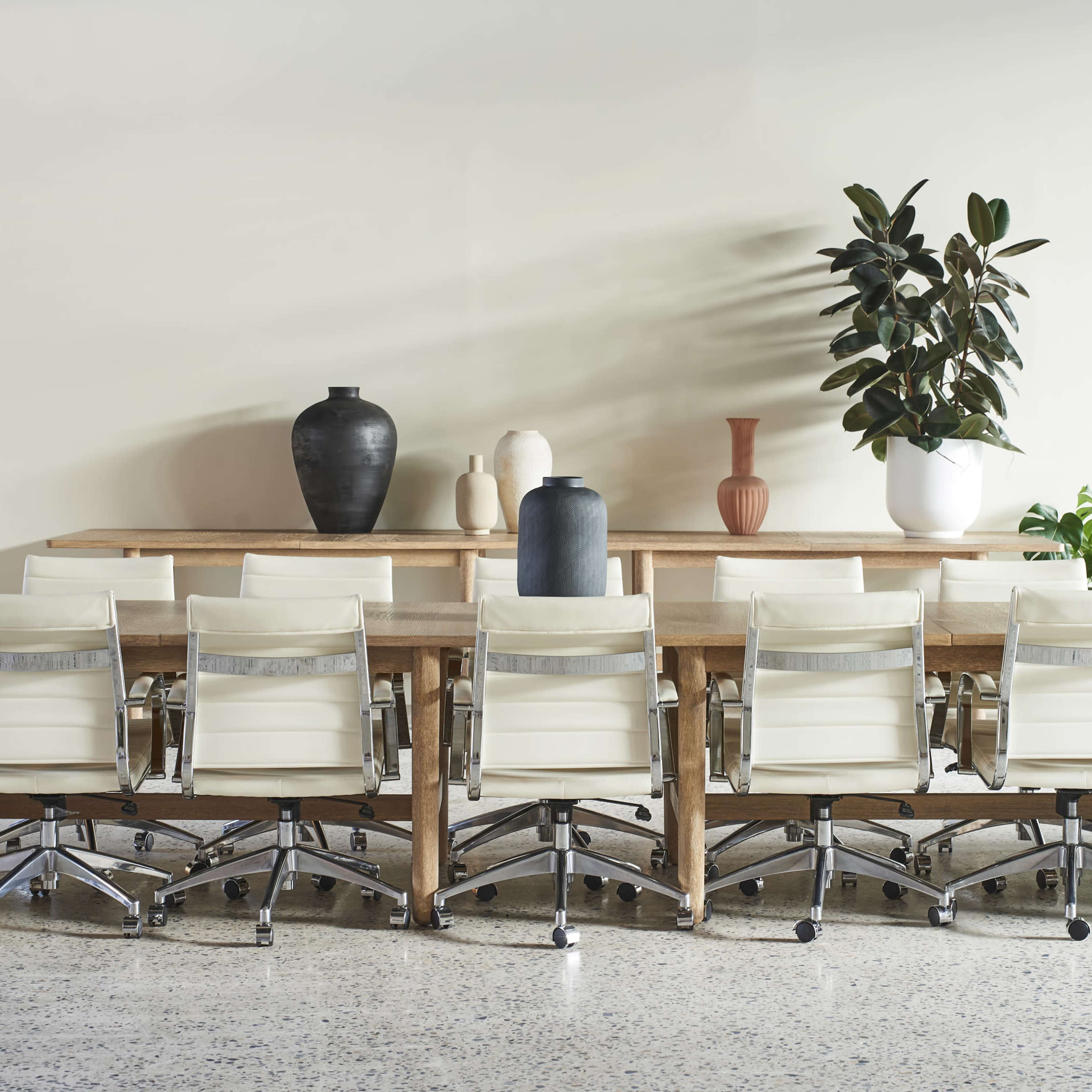 A long wooden conference table with white chairs is arranged in a modern meeting room, featuring decorative vases and a potted plant against a neutral wall.