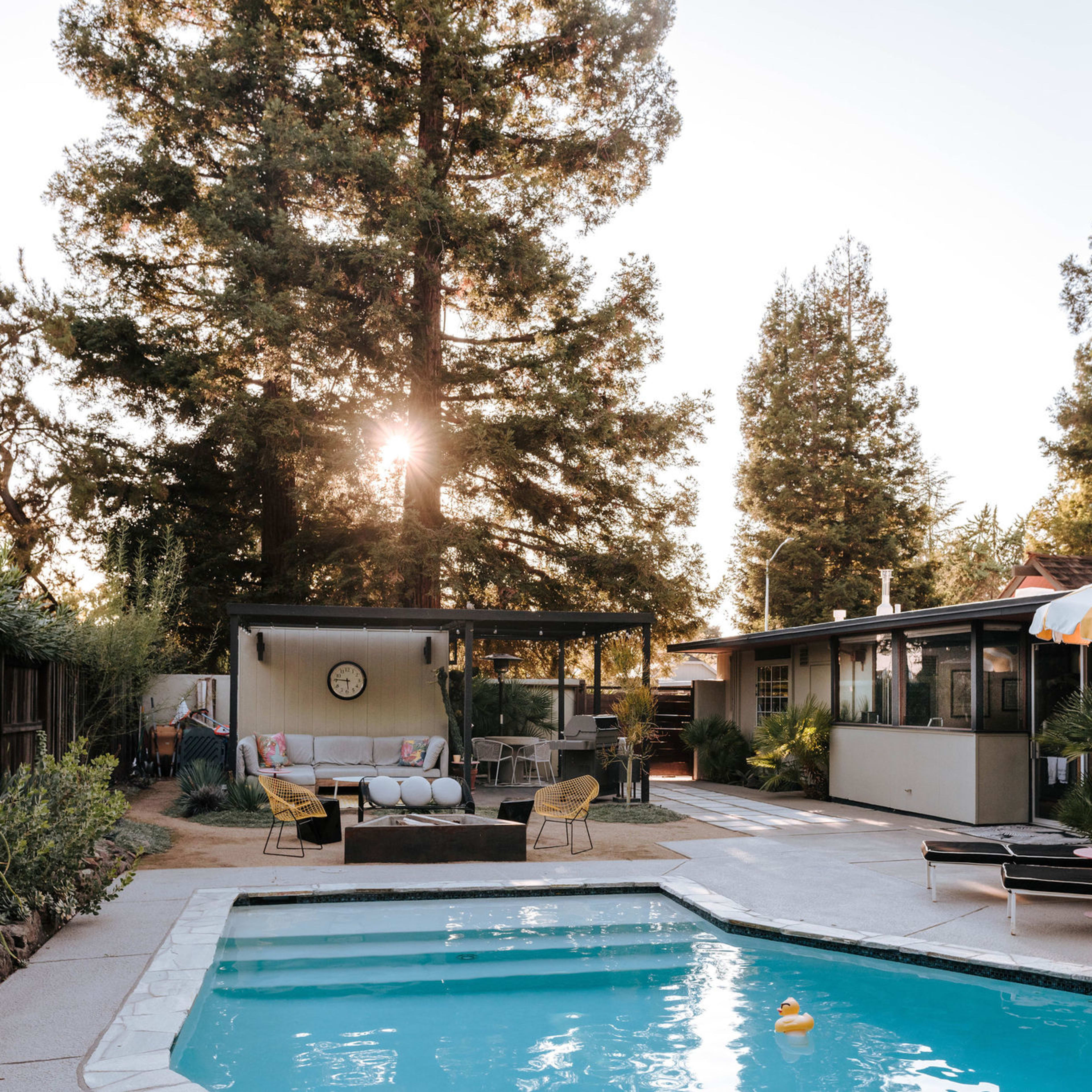 A swimming pool with a yellow-striped umbrella is surrounded by a landscaped yard and a modern outdoor seating area.