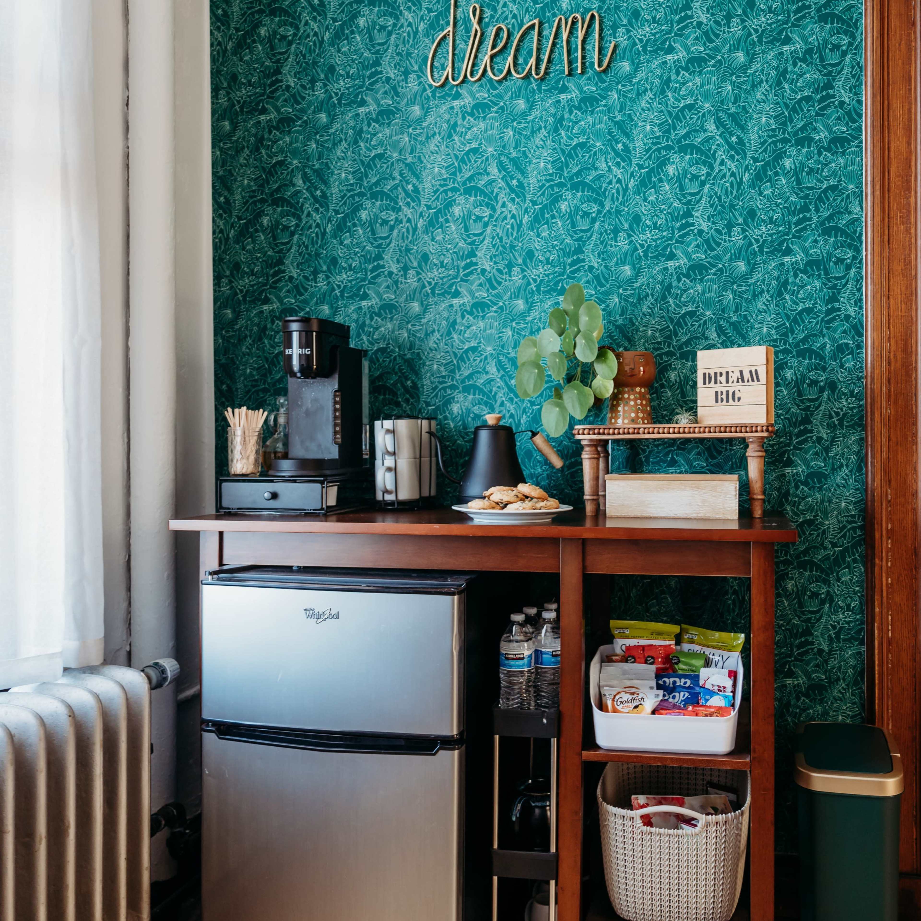 A kitchen corner features a small silver refrigerator, a coffee maker on a wooden table, and various storage baskets against a green floral wallpaper.