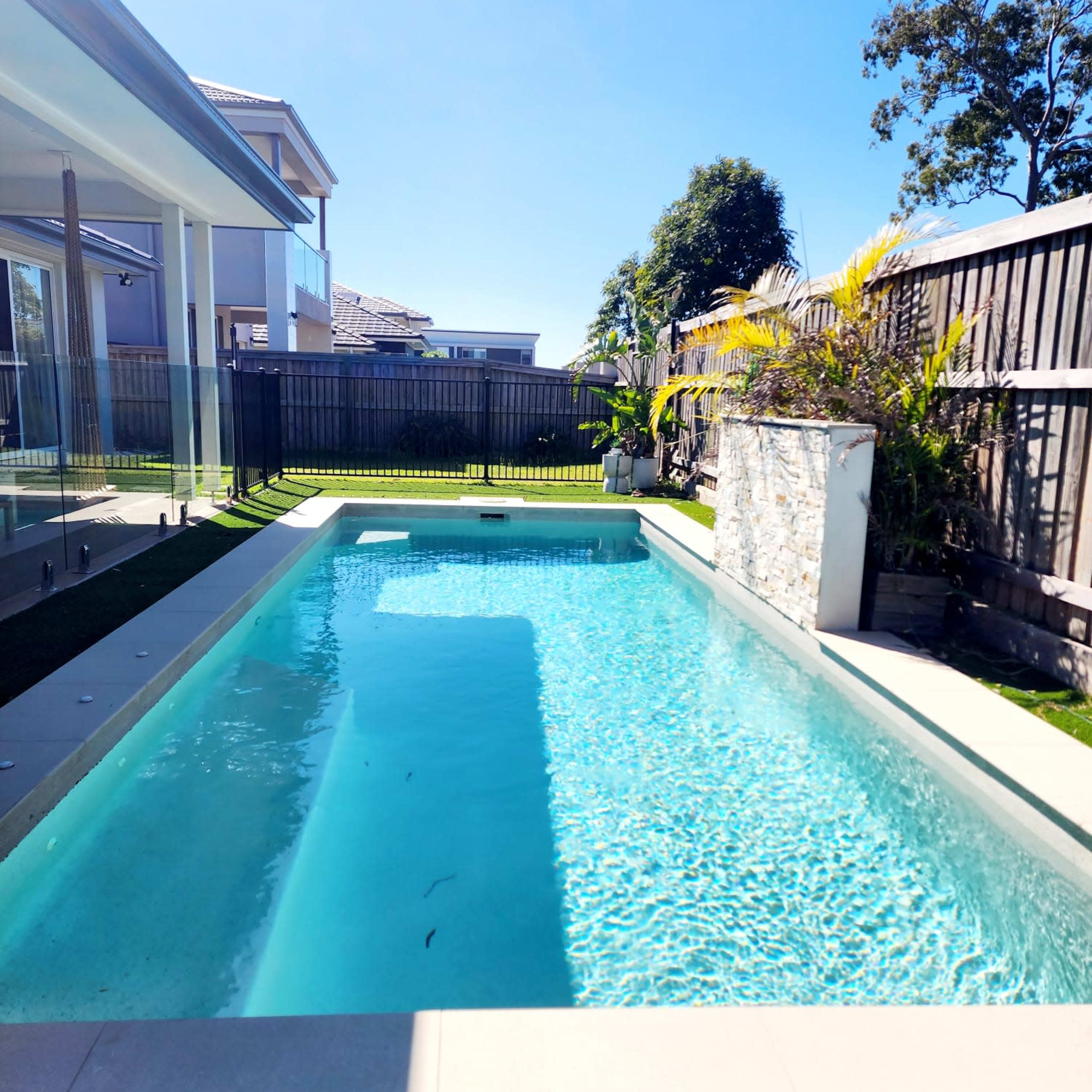 A clear swimming pool surrounded by landscaped greenery and a wooden fence.
