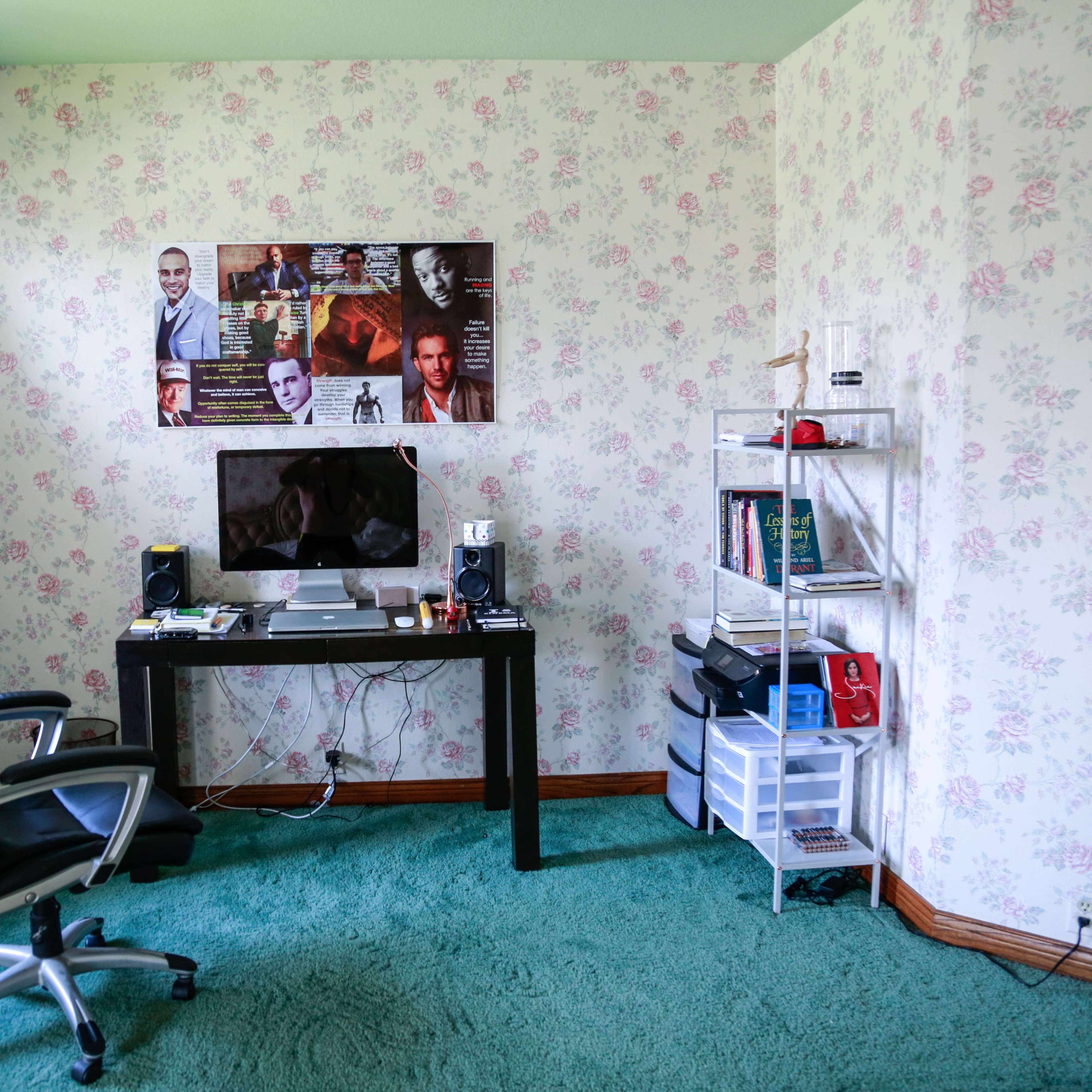 A small home office space with floral wallpaper, a black desk and chair, a computer setup, and a bookshelf against the wall.