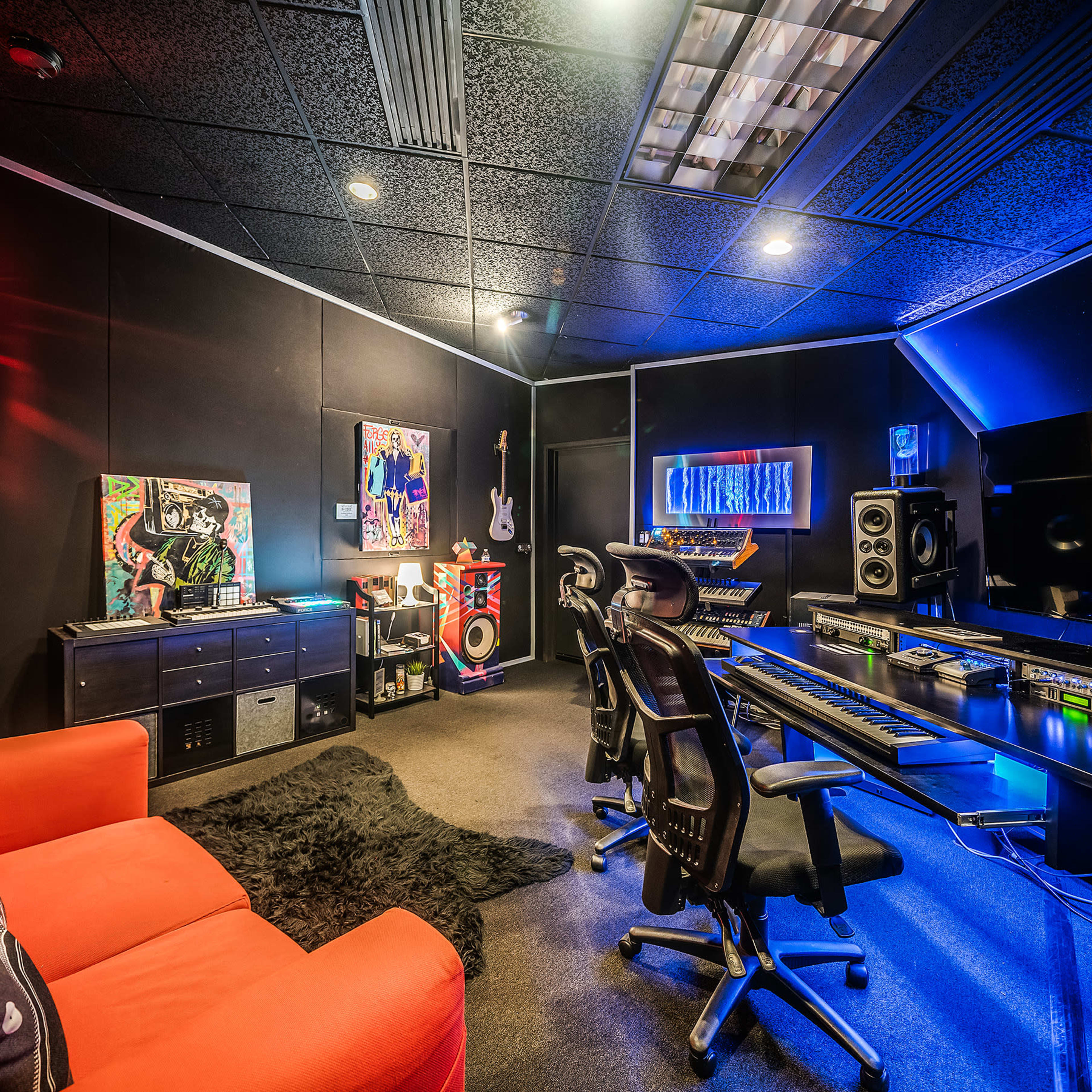 A modern music studio featuring a black wall, a computer desk with audio equipment, a red sofa, and vibrant decorative elements.