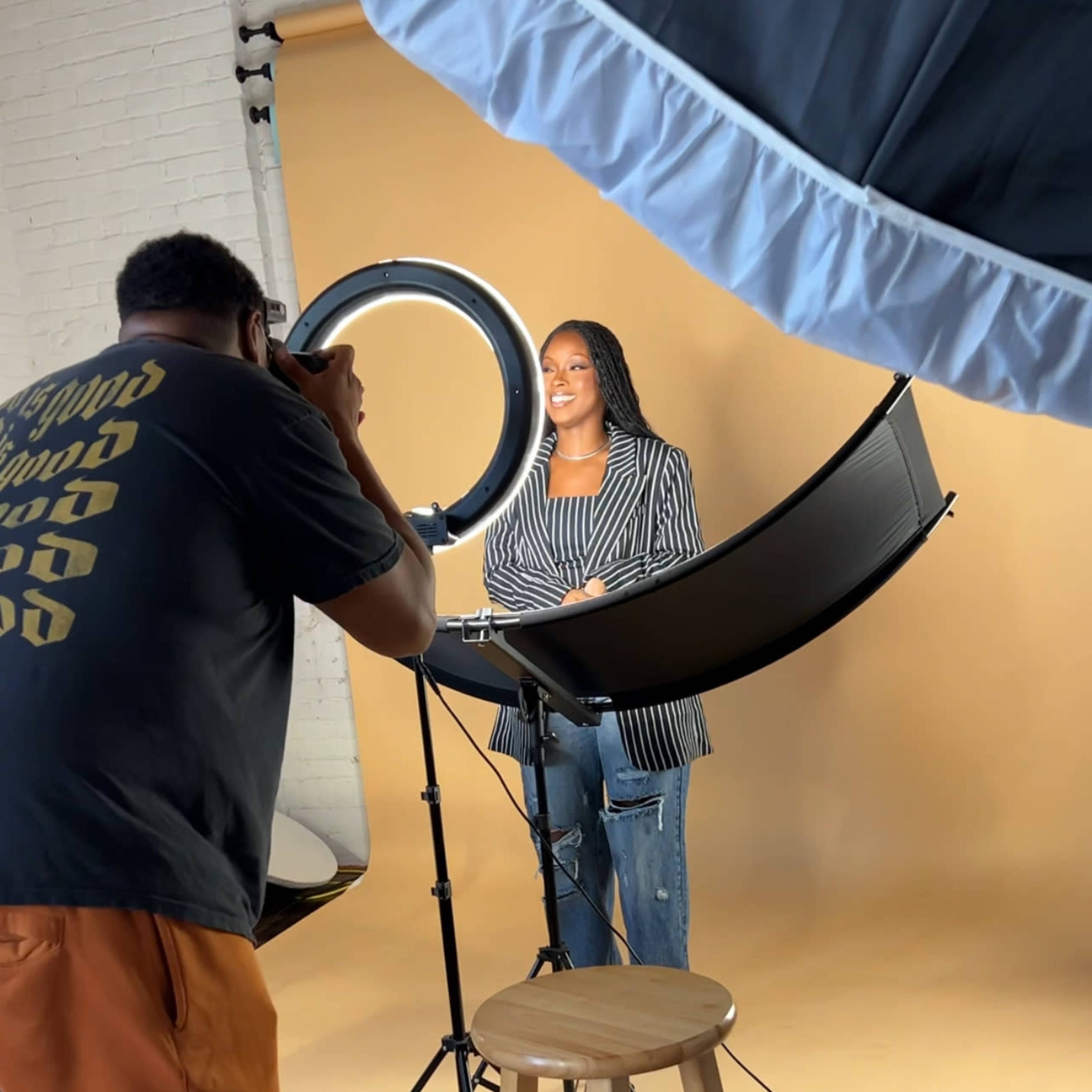 A photographer is taking a portrait of a model standing on a stool in front of a backdrop while using a ring light.