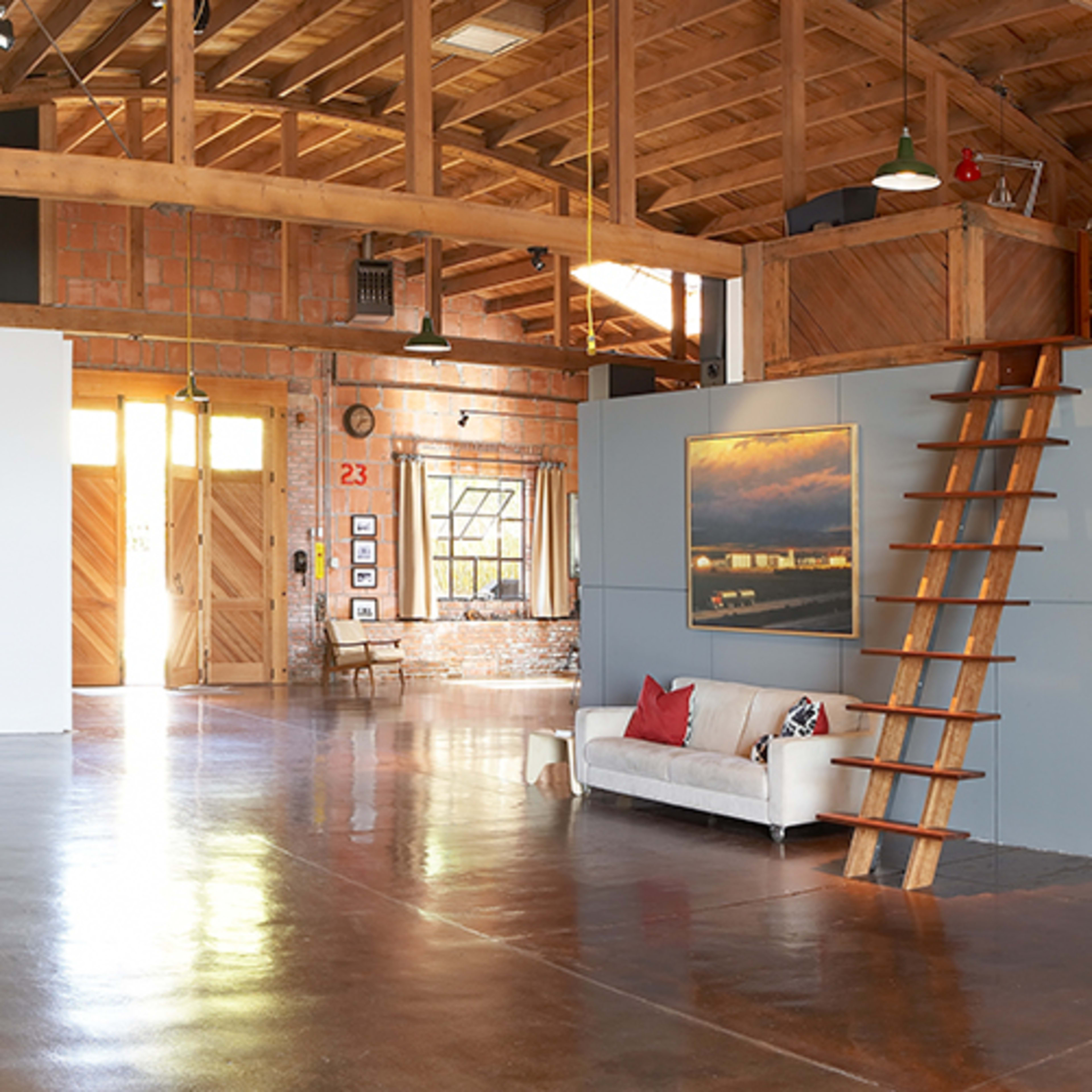 A spacious art gallery with a wooden ceiling, polished concrete floor, and an exhibition wall featuring a large photo alongside a staircase leading to a loft area.