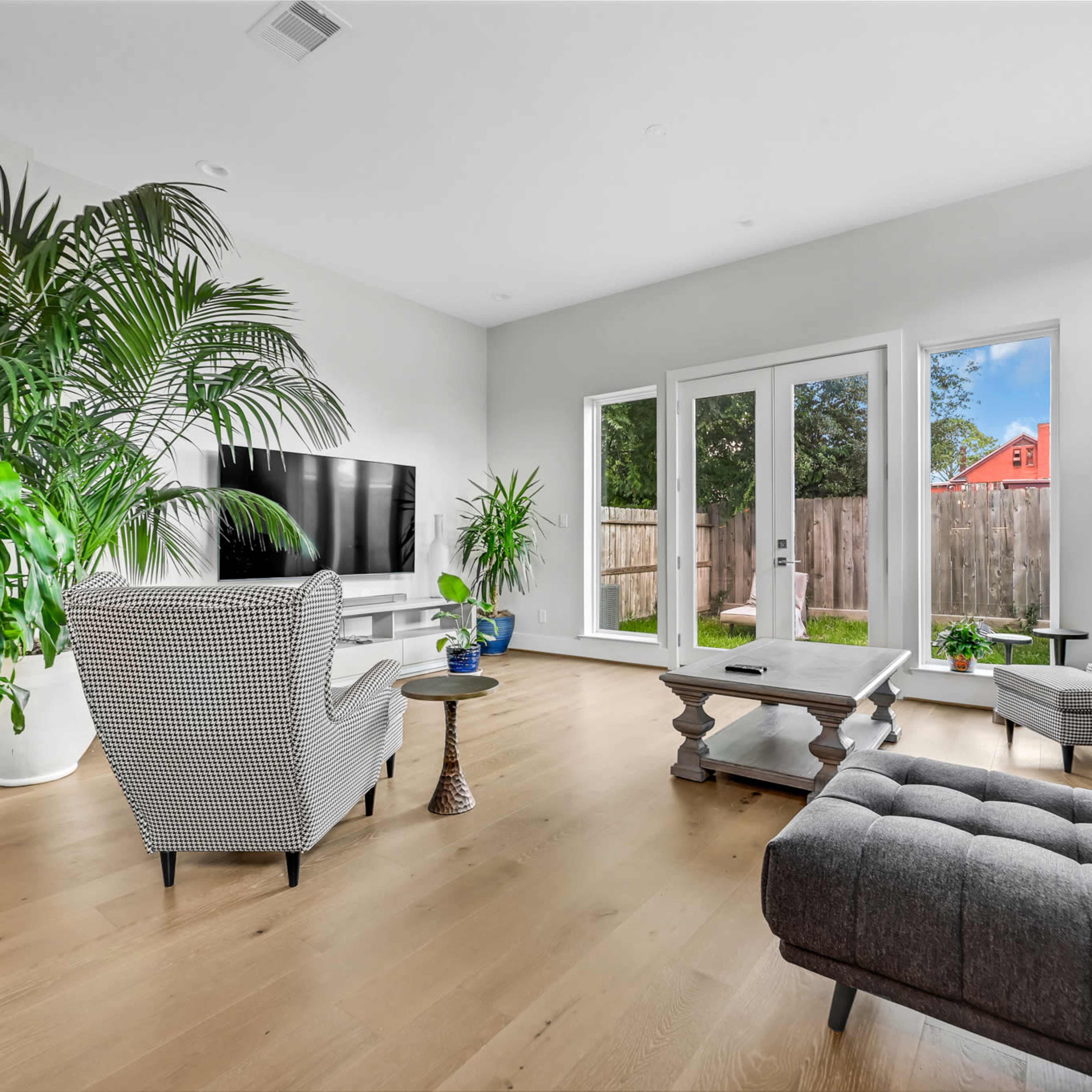 The living room features a large television, modern furniture, and several potted plants by large windows that overlook a fenced backyard.