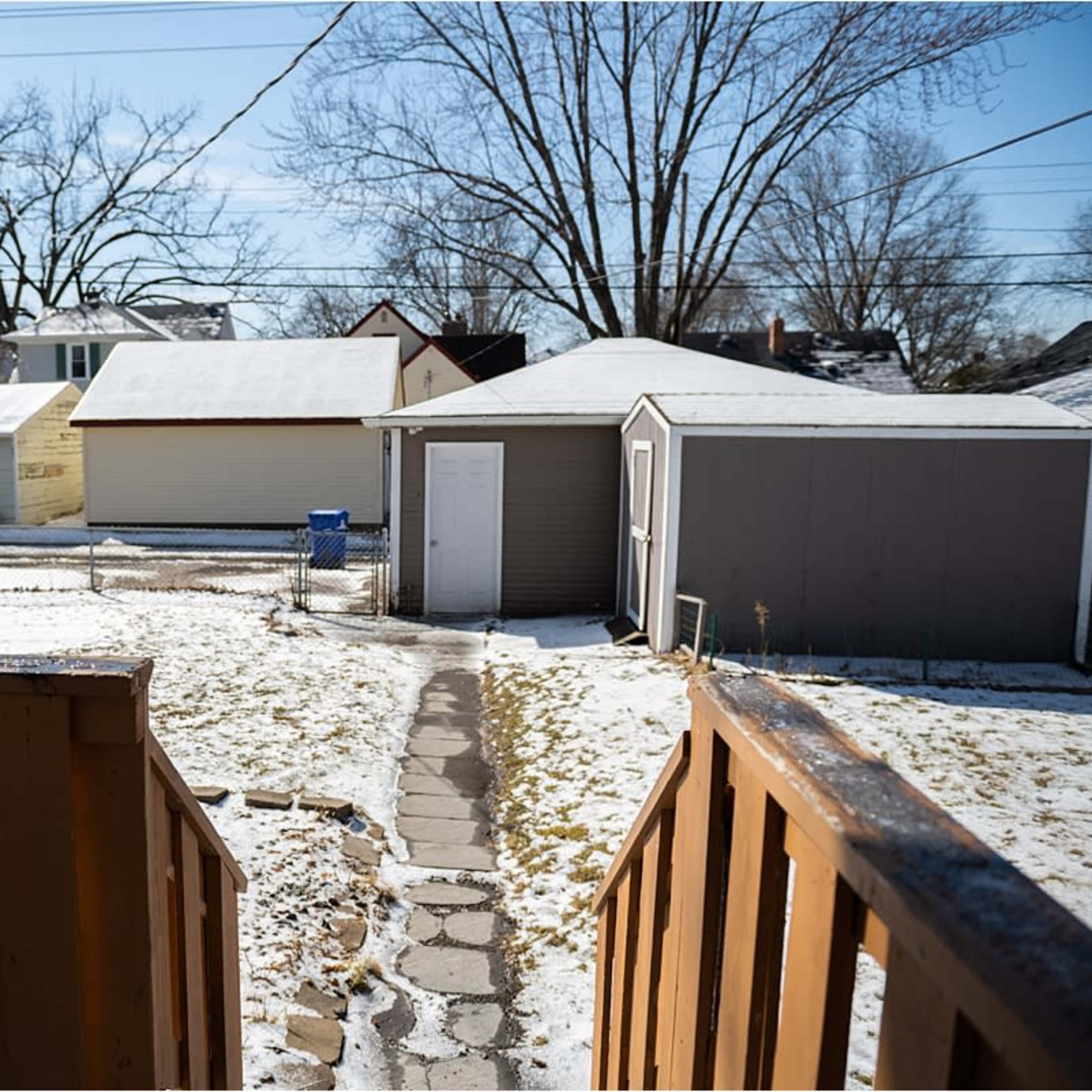 A backyard scene shows a pathway leading to a gray shed, surrounded by snow-covered ground and residential buildings in the background.