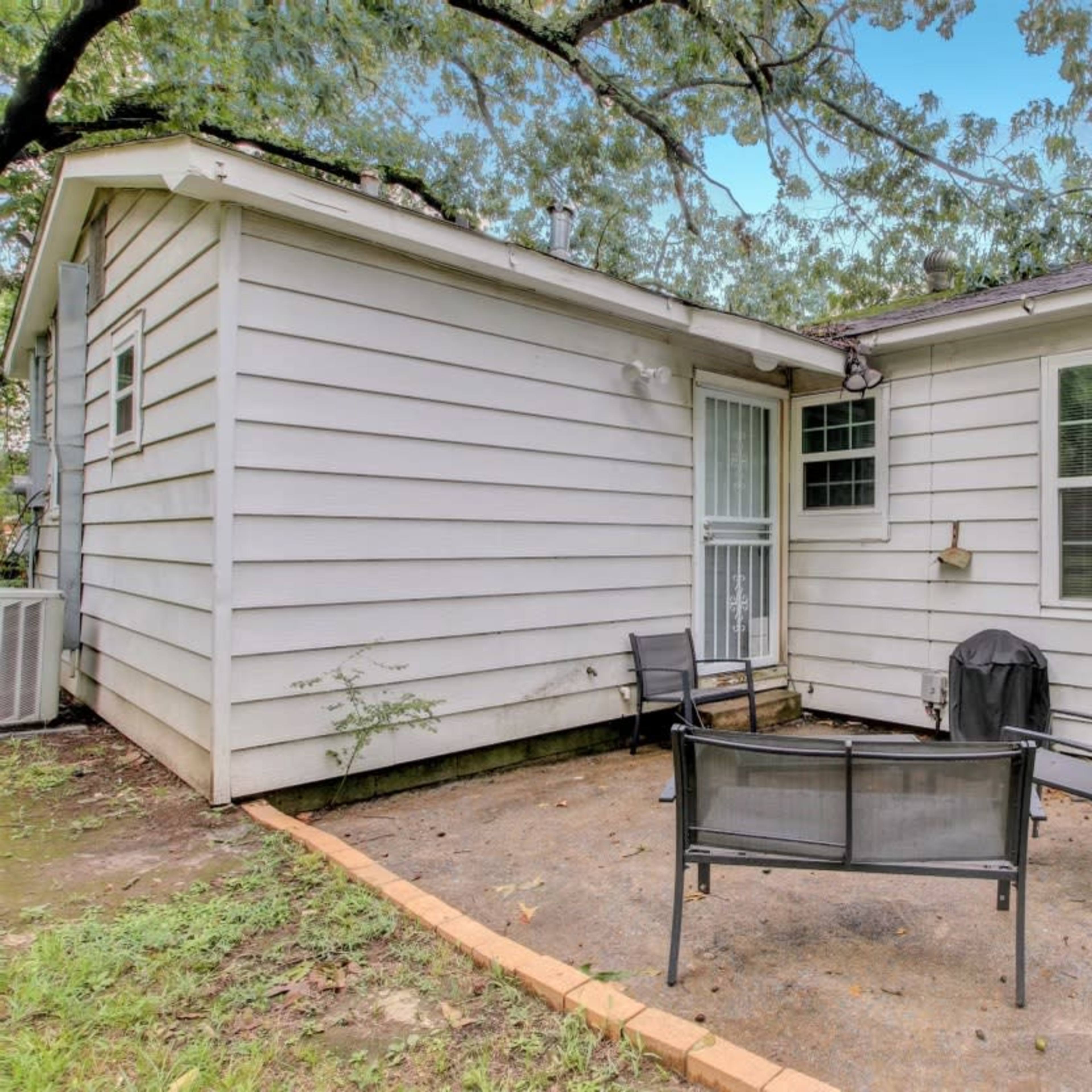 A residential exterior area with a small patio featuring four chairs and a grill beside a single-story house.