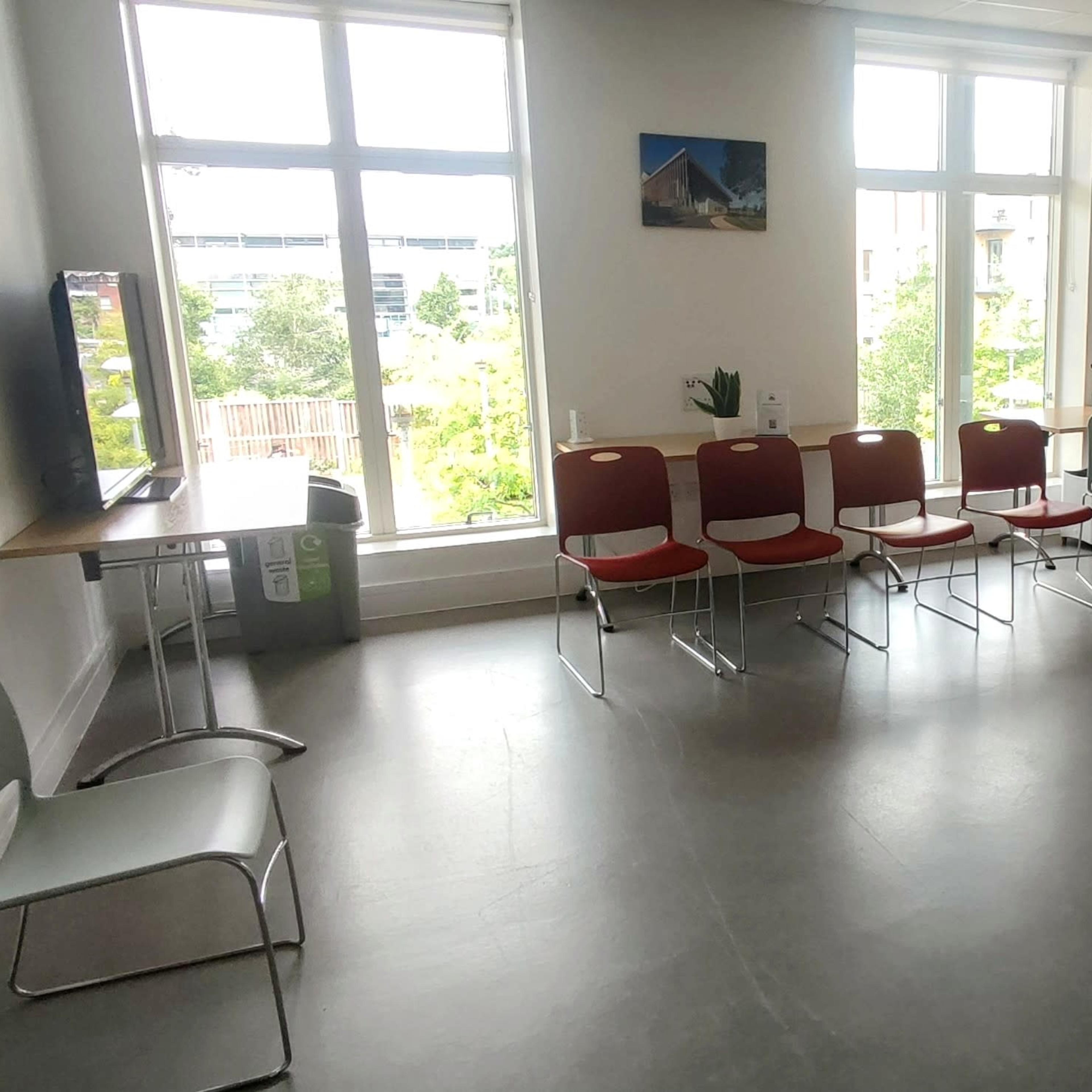 A meeting room features a circular arrangement of red and light blue chairs around a small table, with large windows allowing natural light to enter.