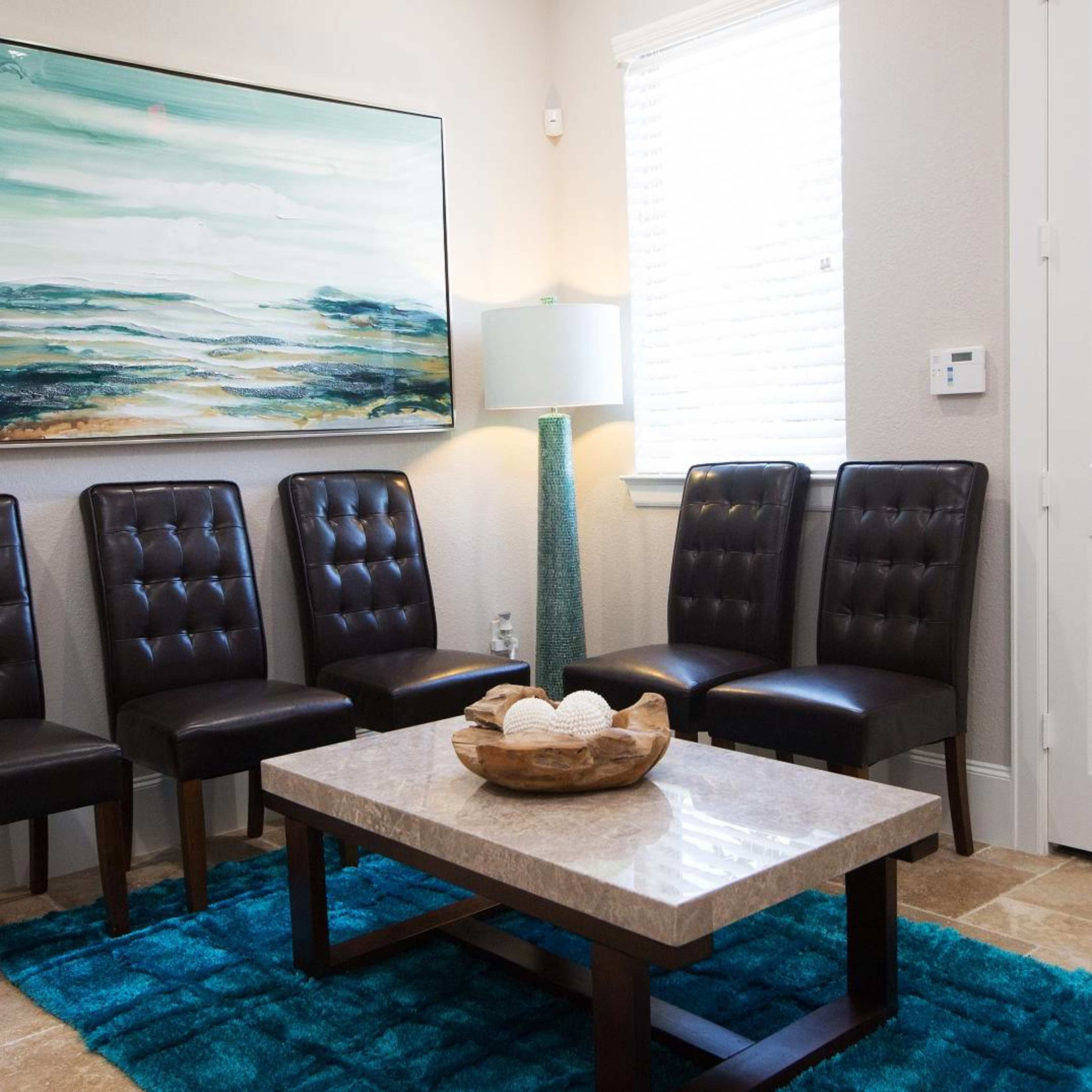 A waiting area with six black leather chairs arranged around a marble coffee table on a blue rug, accompanied by an abstract painting and a lamp.