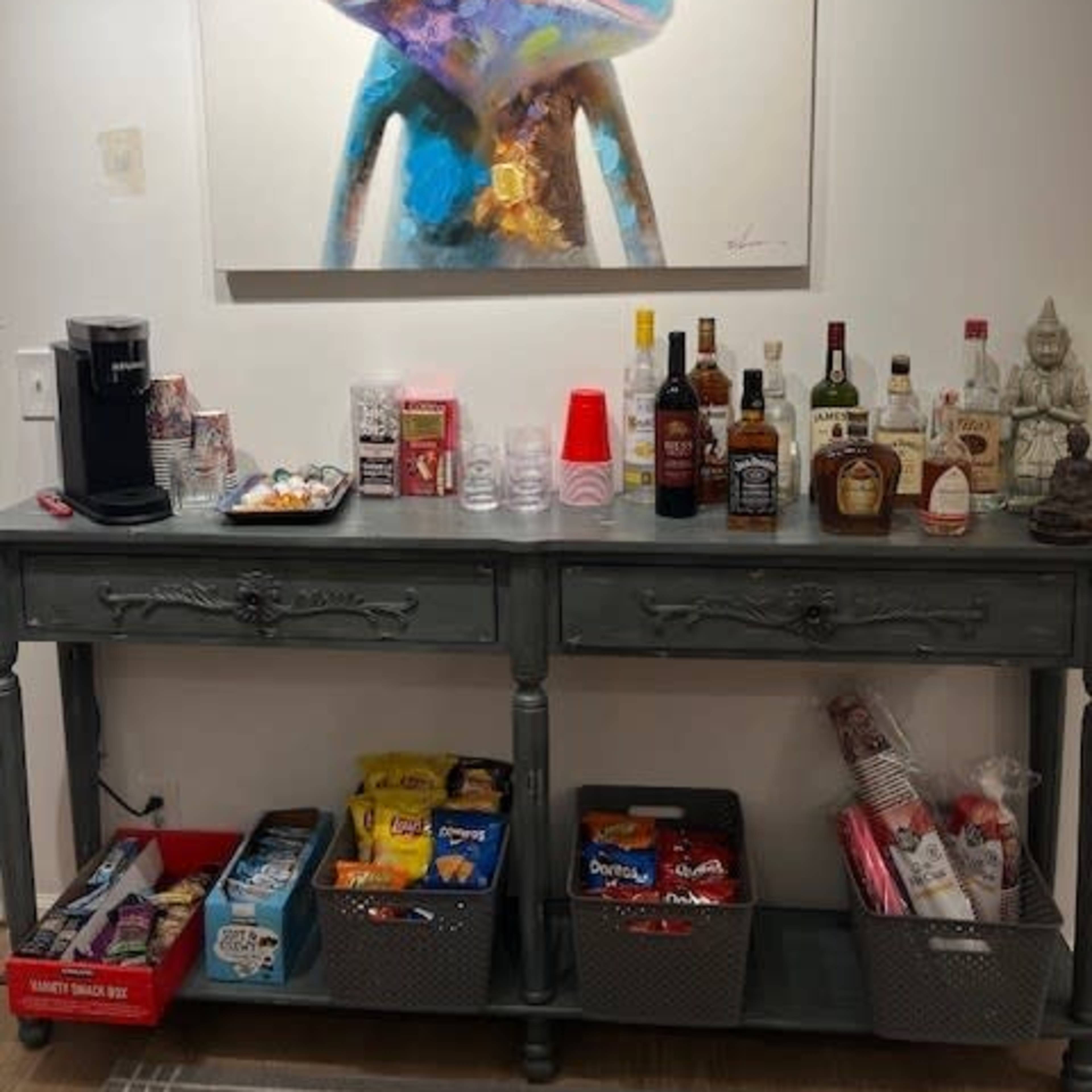 A table holds various snacks and drinks beneath a colorful frog painting wearing large glasses.