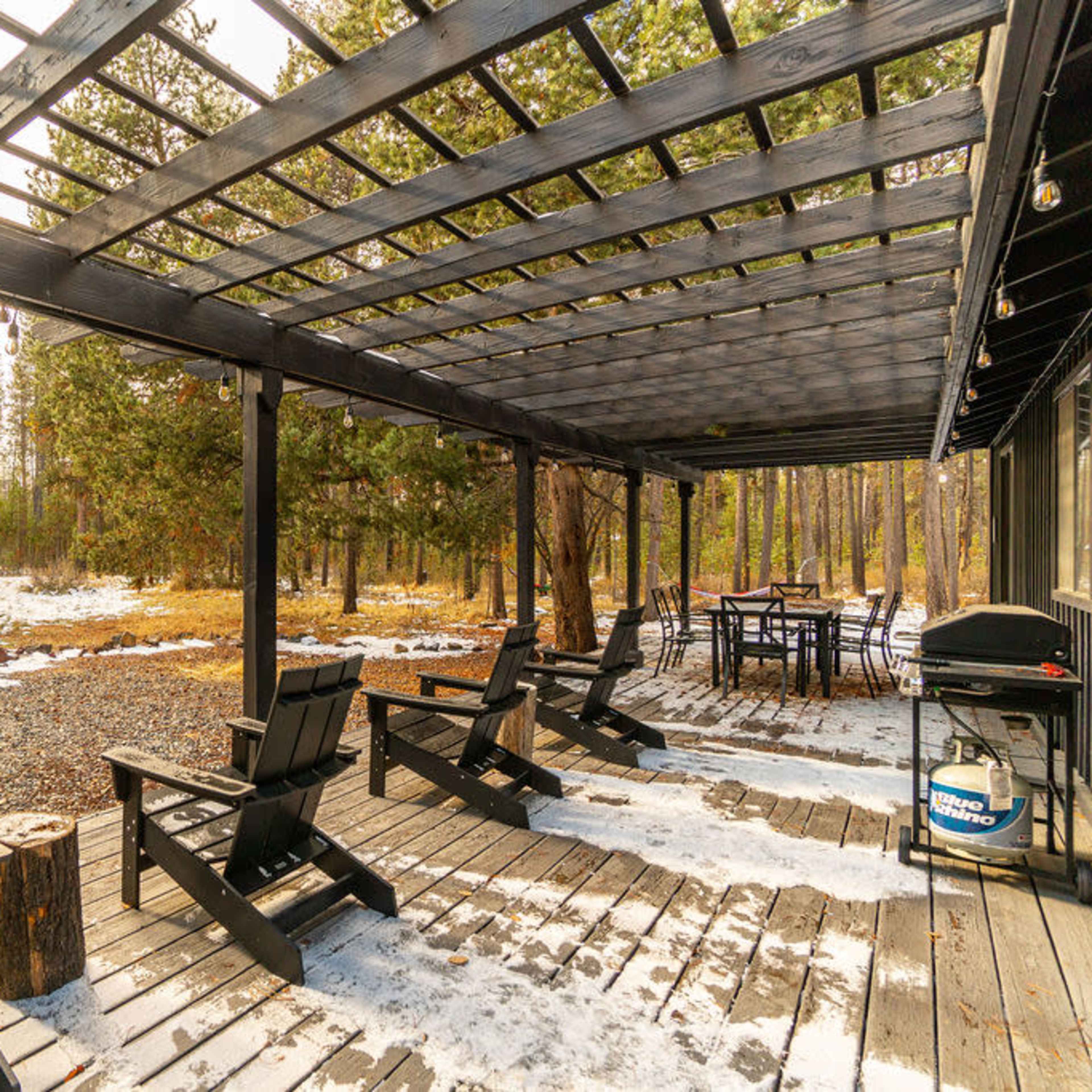 The scene features a black wooden deck with several Adirondack chairs overlooking a snowy landscape filled with trees.