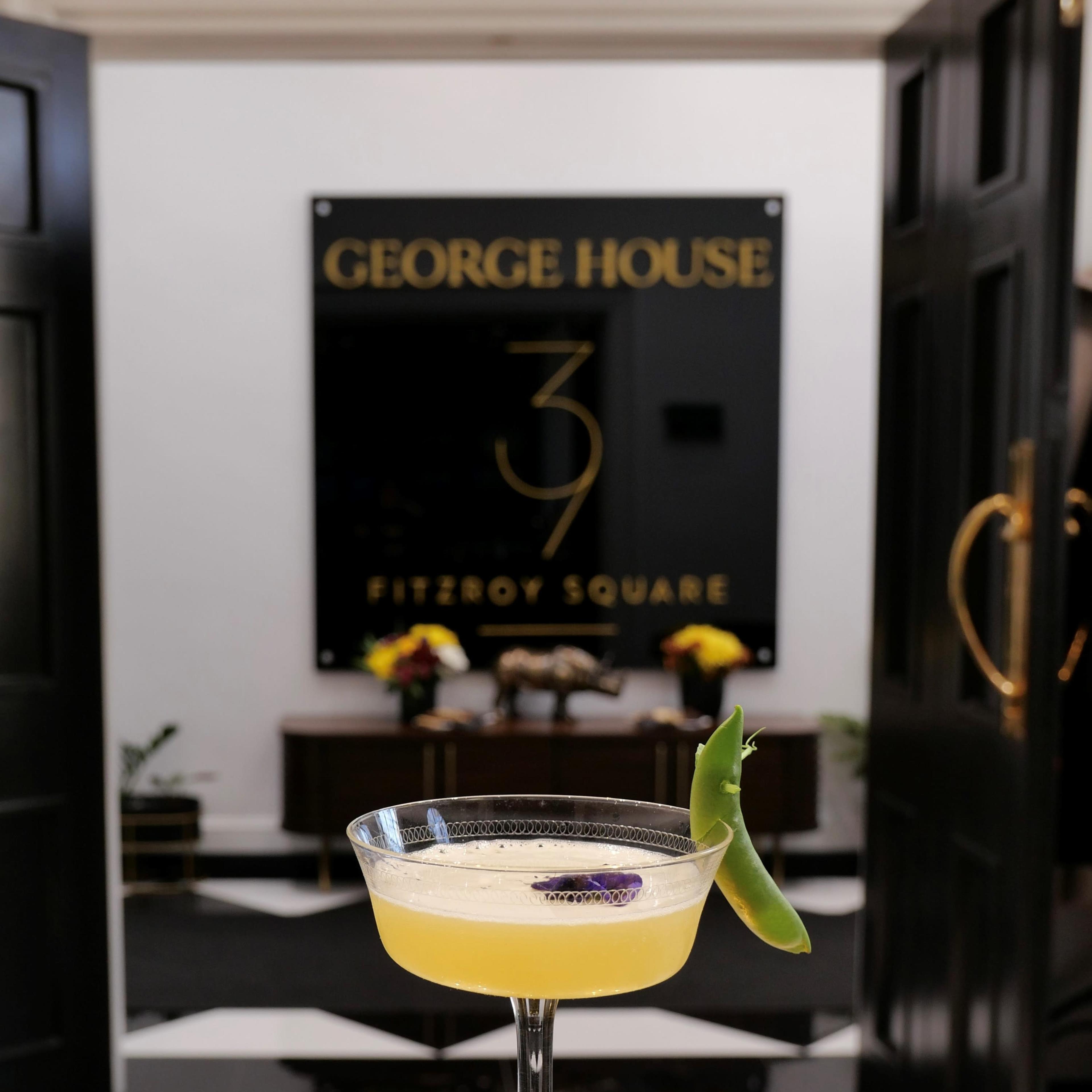 A cocktail is served on a table in front of a sign that displays "George House 39 Fitzroy Square."