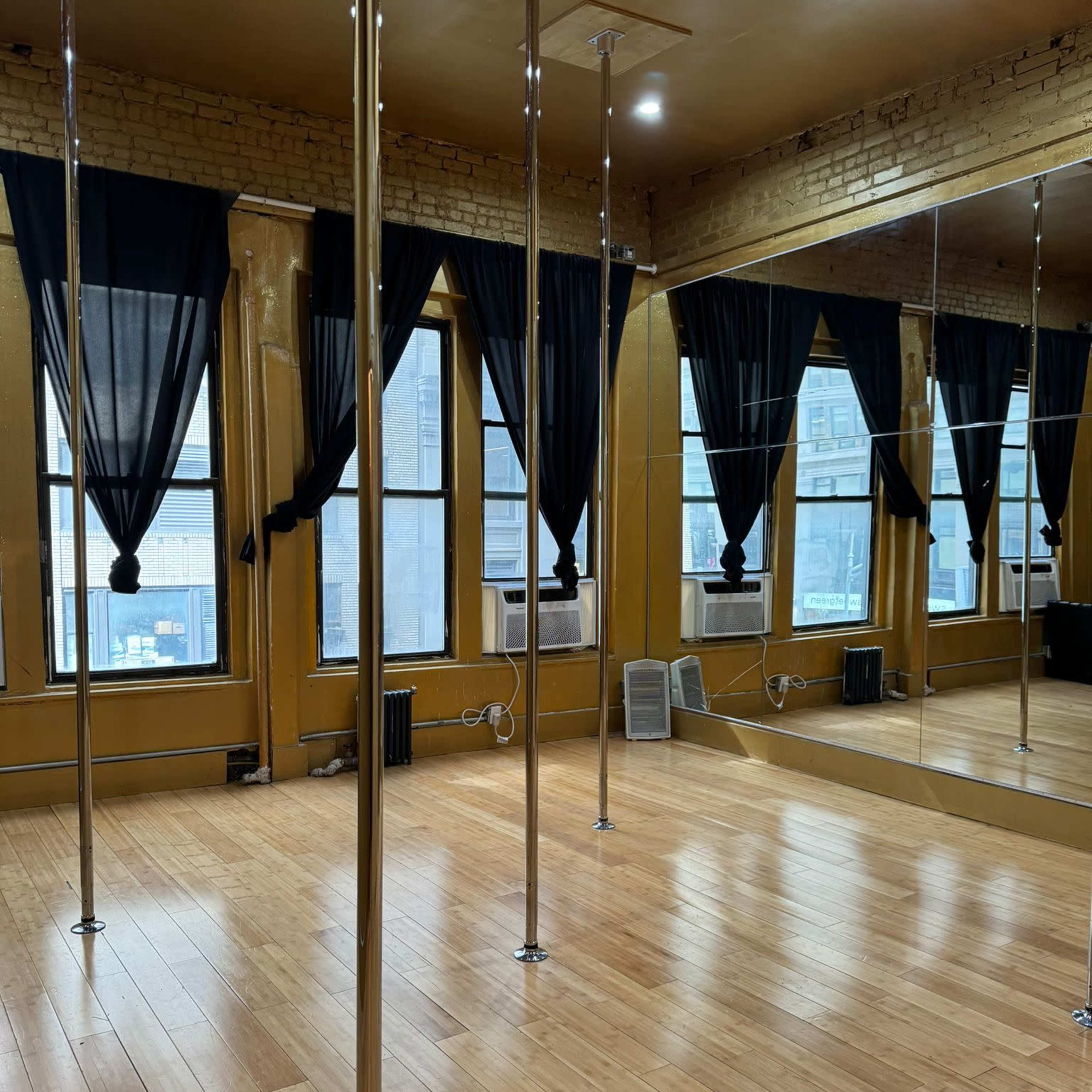 A dance studio with wooden floors, several polished metal poles, large windows draped with black curtains, and a mirrored wall.