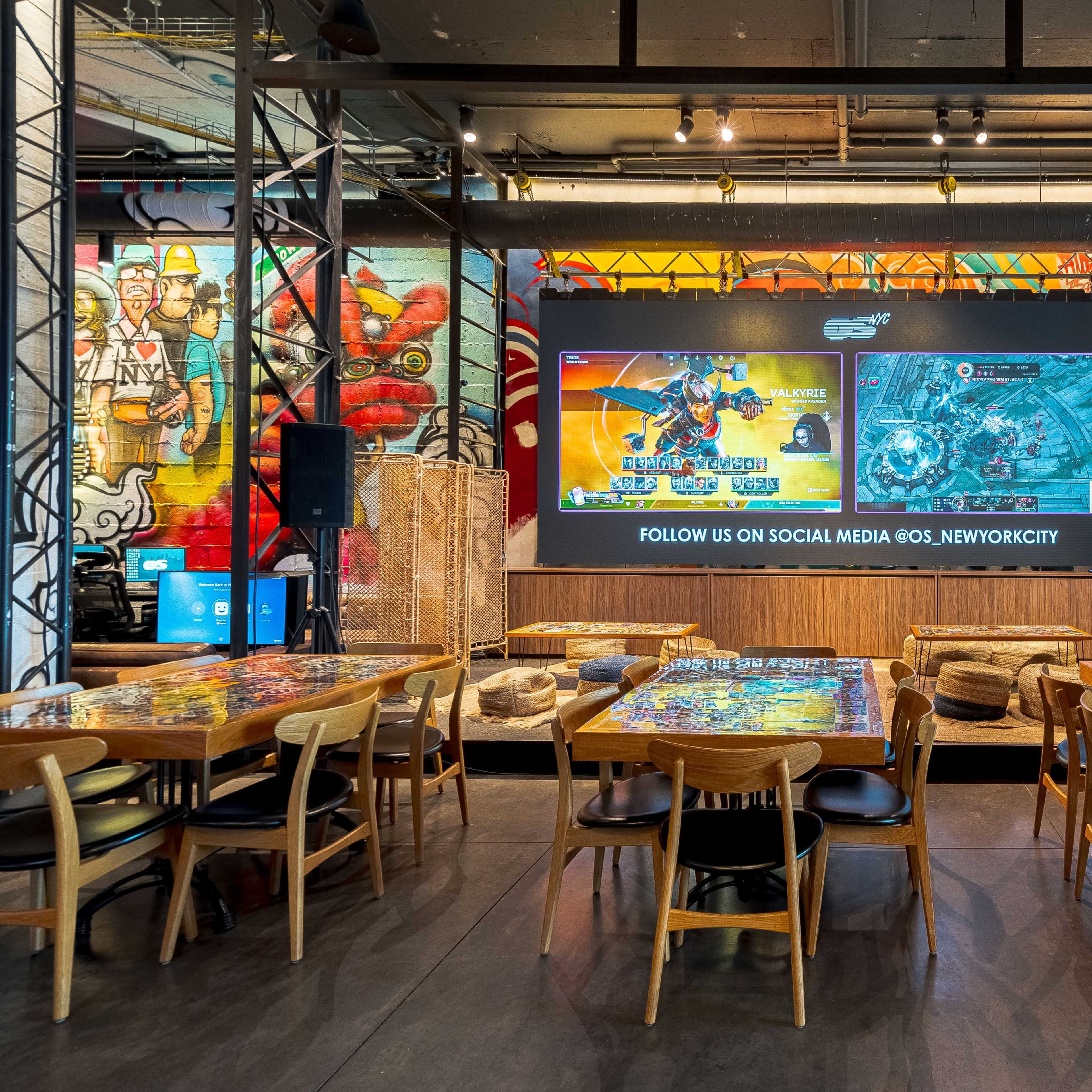 An entertainment venue with large screens displaying video game content, seating areas with wooden tables, and colorful murals on the walls.