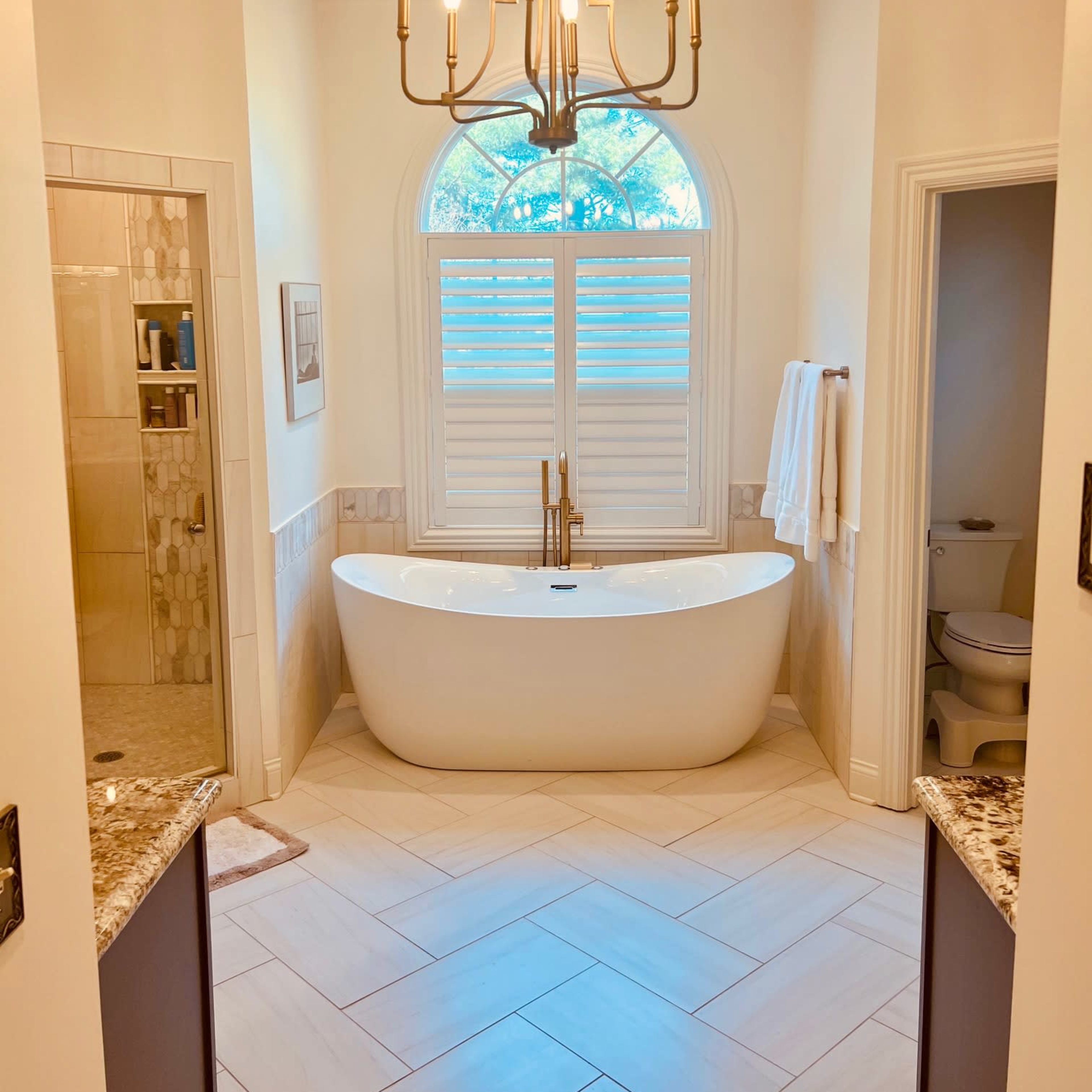 A spacious bathroom featuring a freestanding white bathtub, a large window with shutters, and a chandelier above.