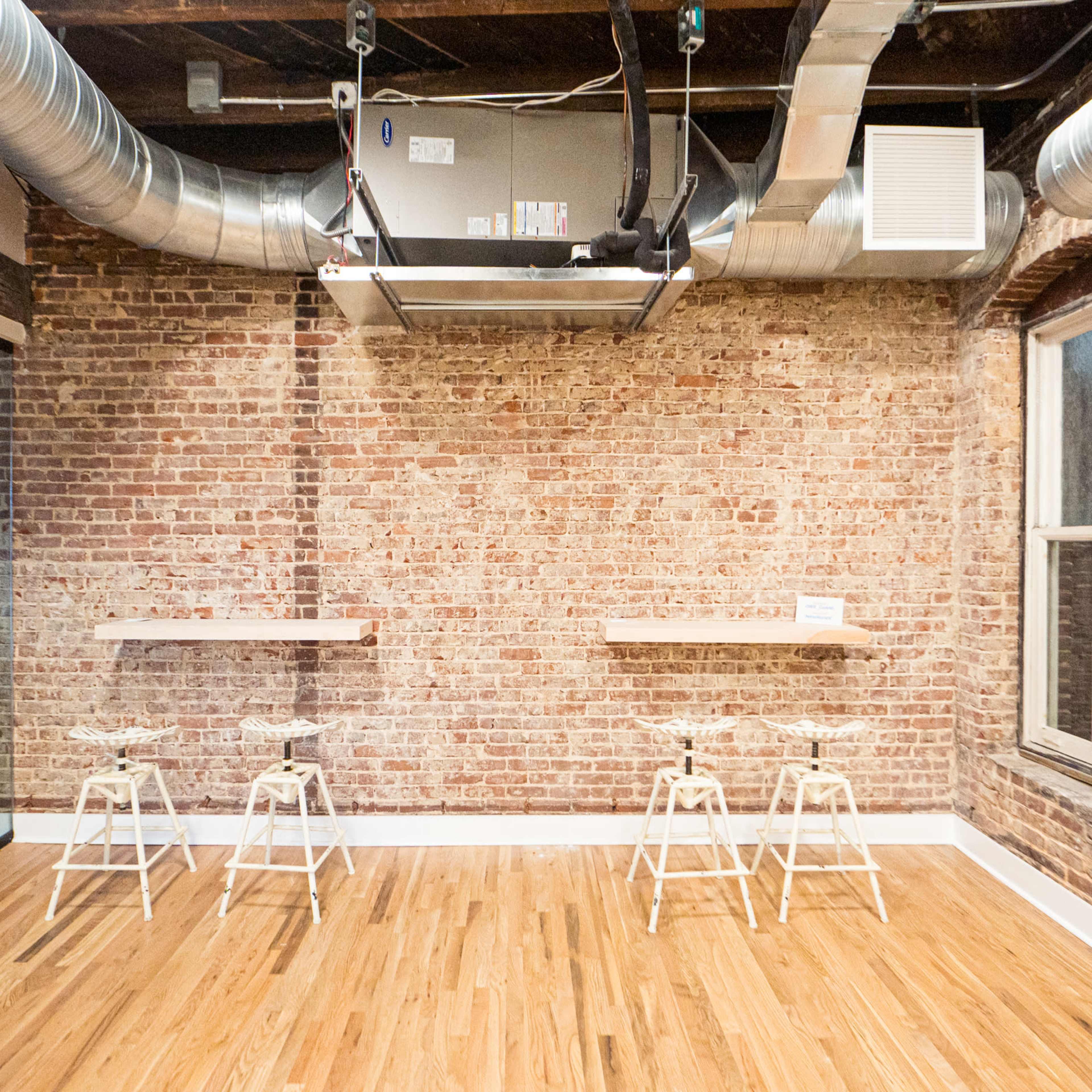 An industrial-style office space with exposed brick walls, wooden flooring, and three high stools positioned near a bar-height counter.