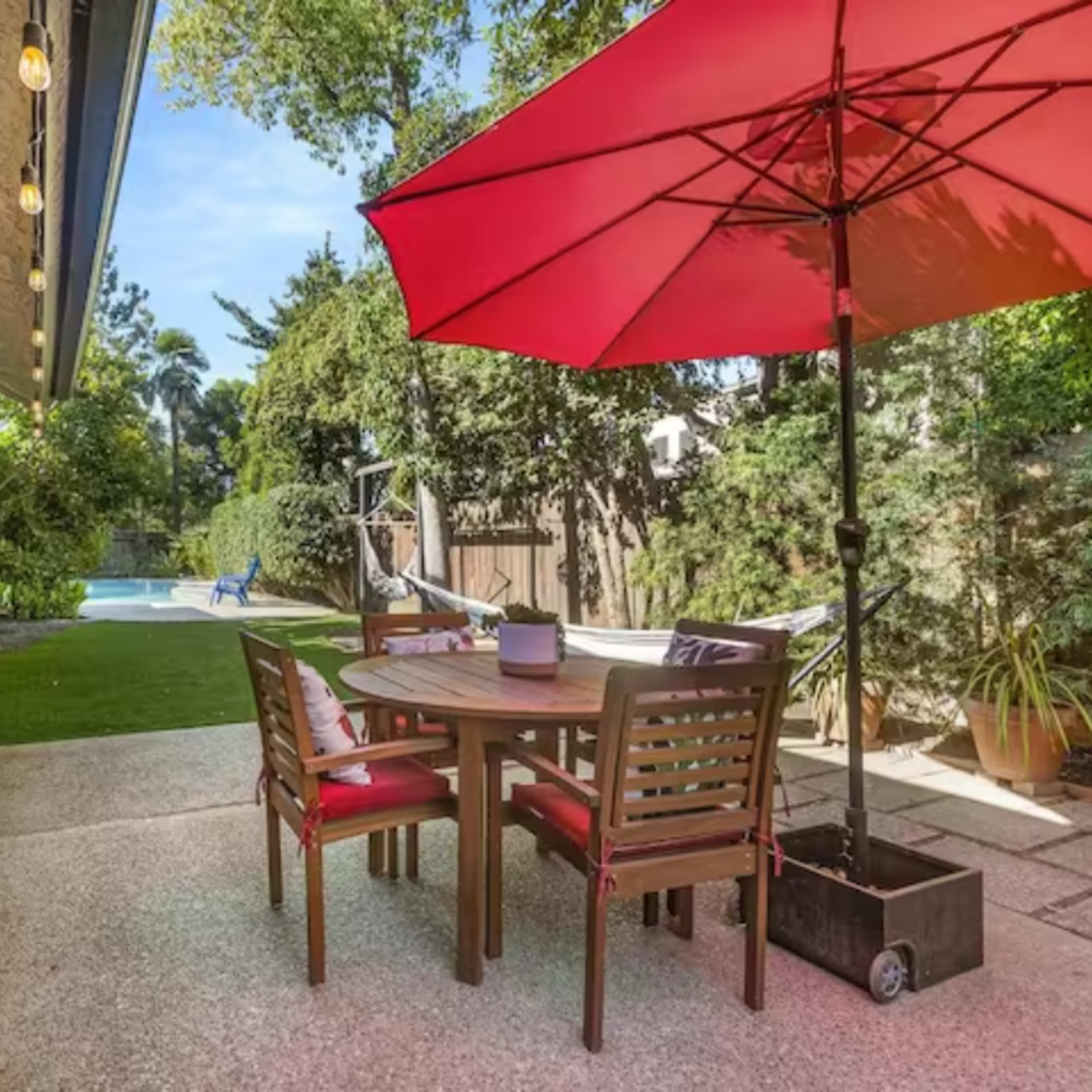 A patio area features a round wooden dining table with chairs, shaded by a red umbrella, overlooking a grassy yard and swimming pool.