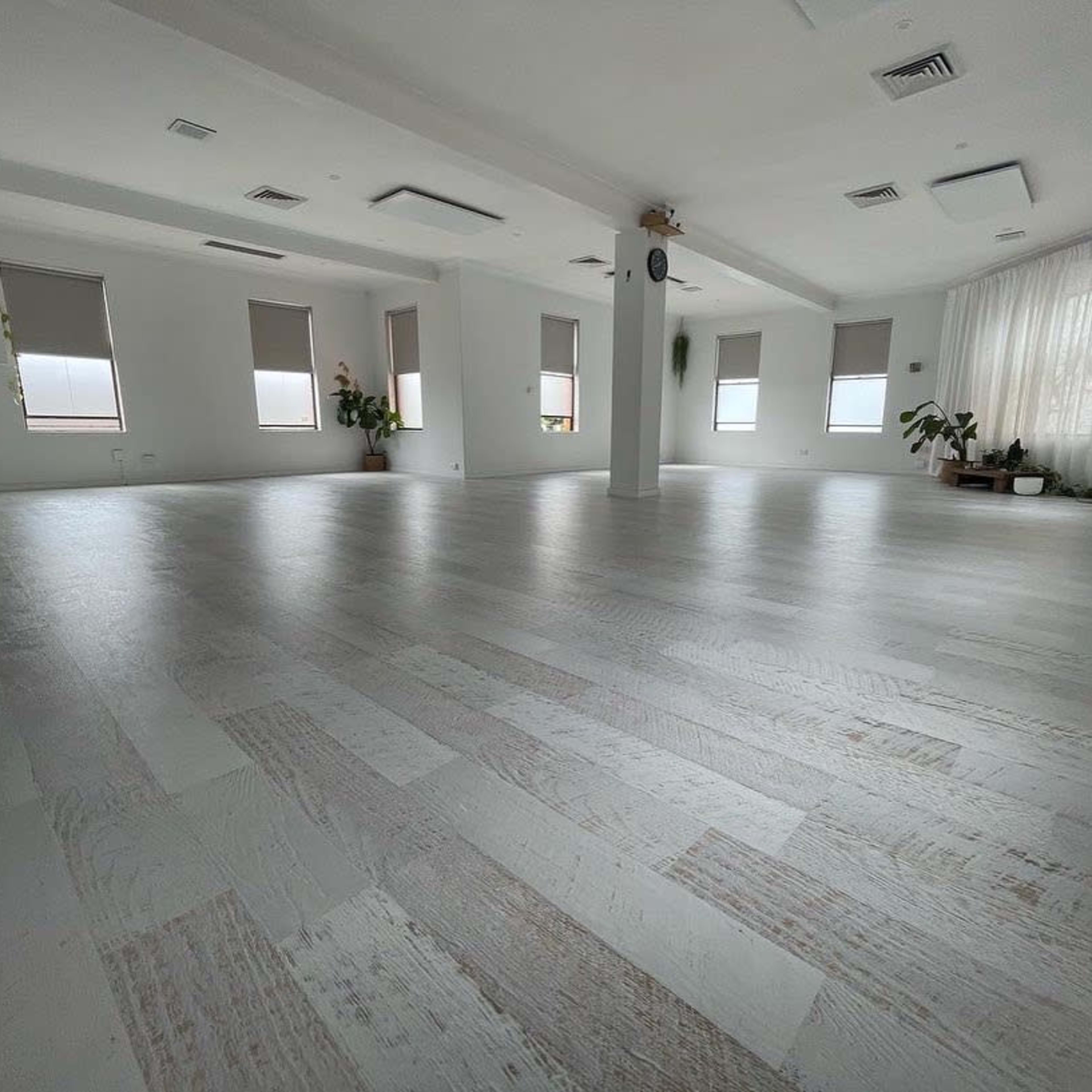 A spacious, well-lit room with large windows, minimal furniture, and plants along the walls.