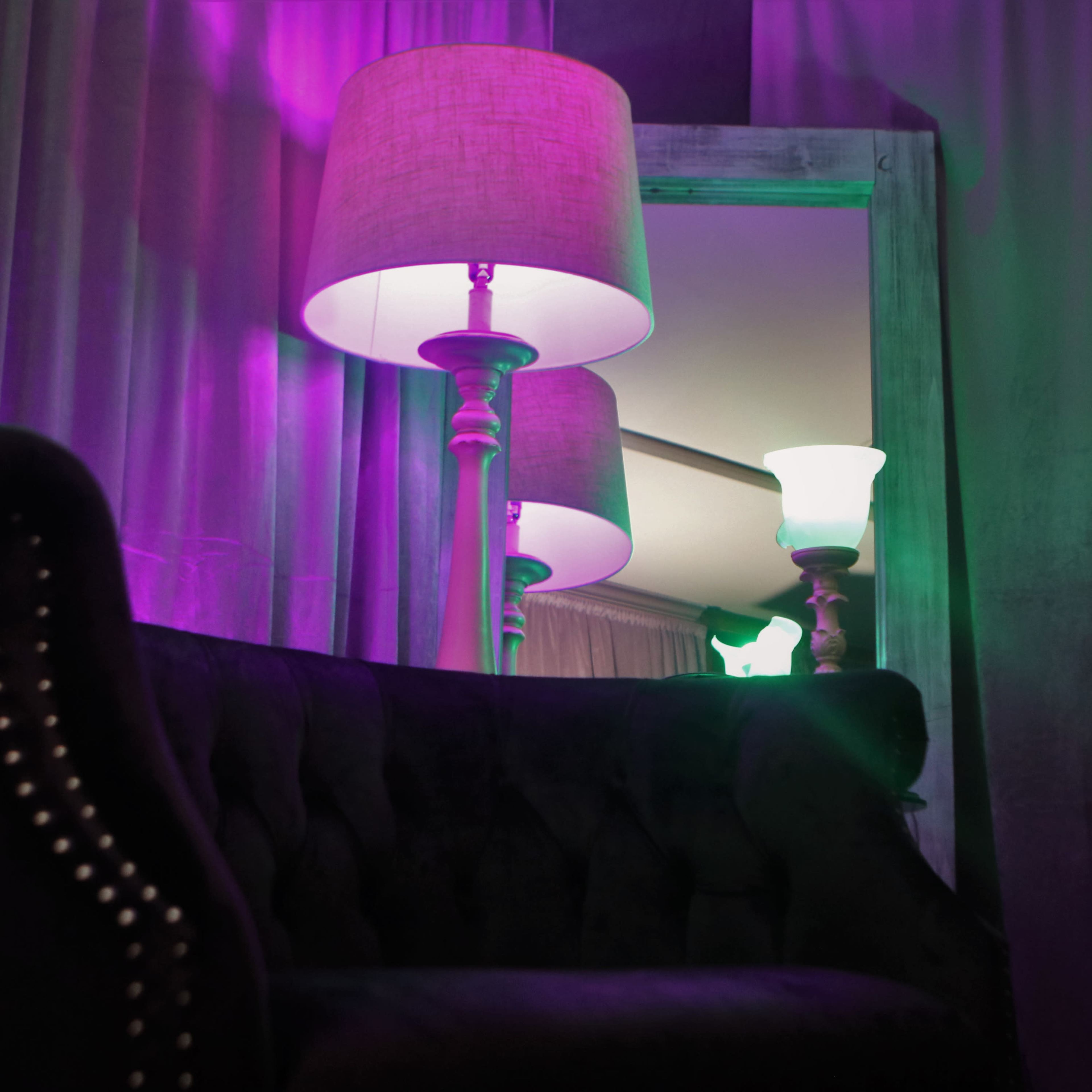 A dark sofa sits in front of a mirror reflecting two lamps, one purple and one green, casting colored light on the curtains.