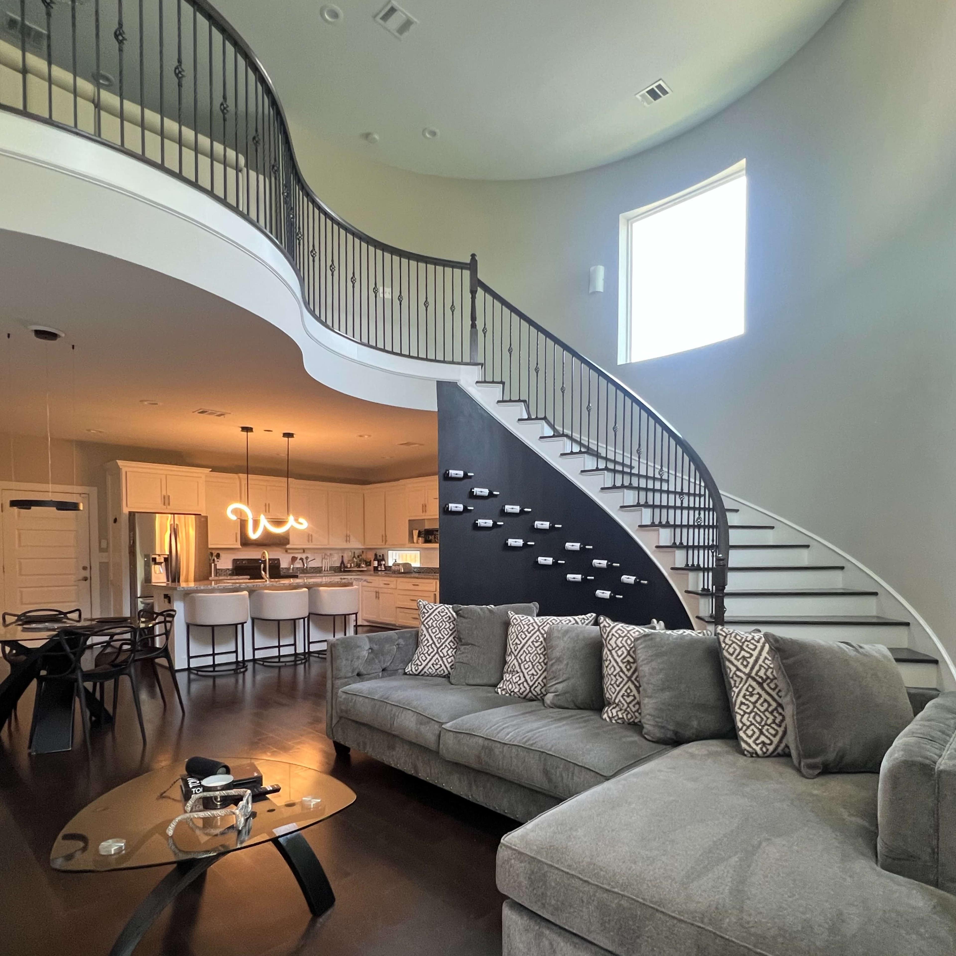 A modern living room with a large gray sectional sofa, a coffee table, and a sweeping staircase that leads to an upper floor.