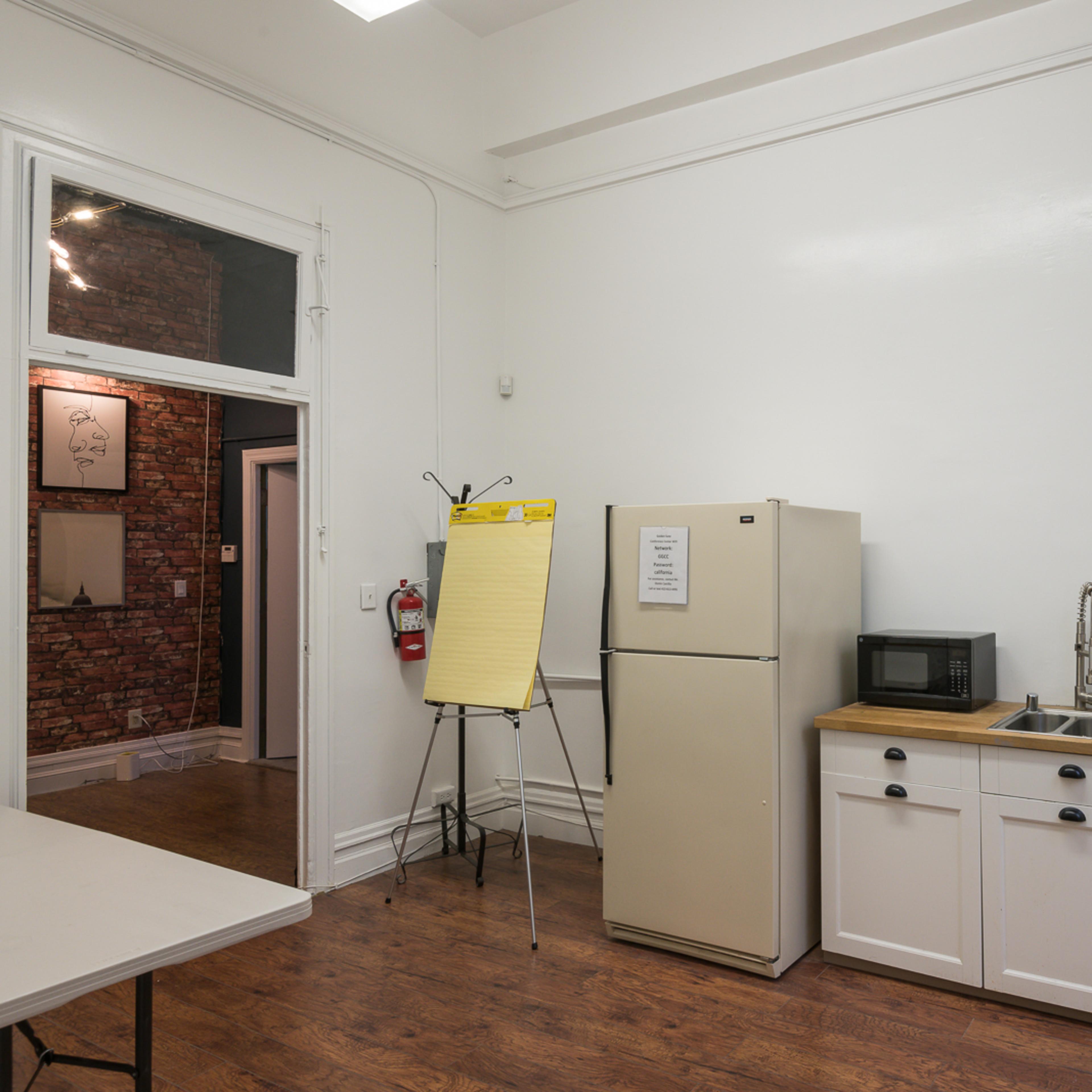 A small kitchen area features a refrigerator, microwave, sink, and a countertop in a well-lit room with wooden flooring.