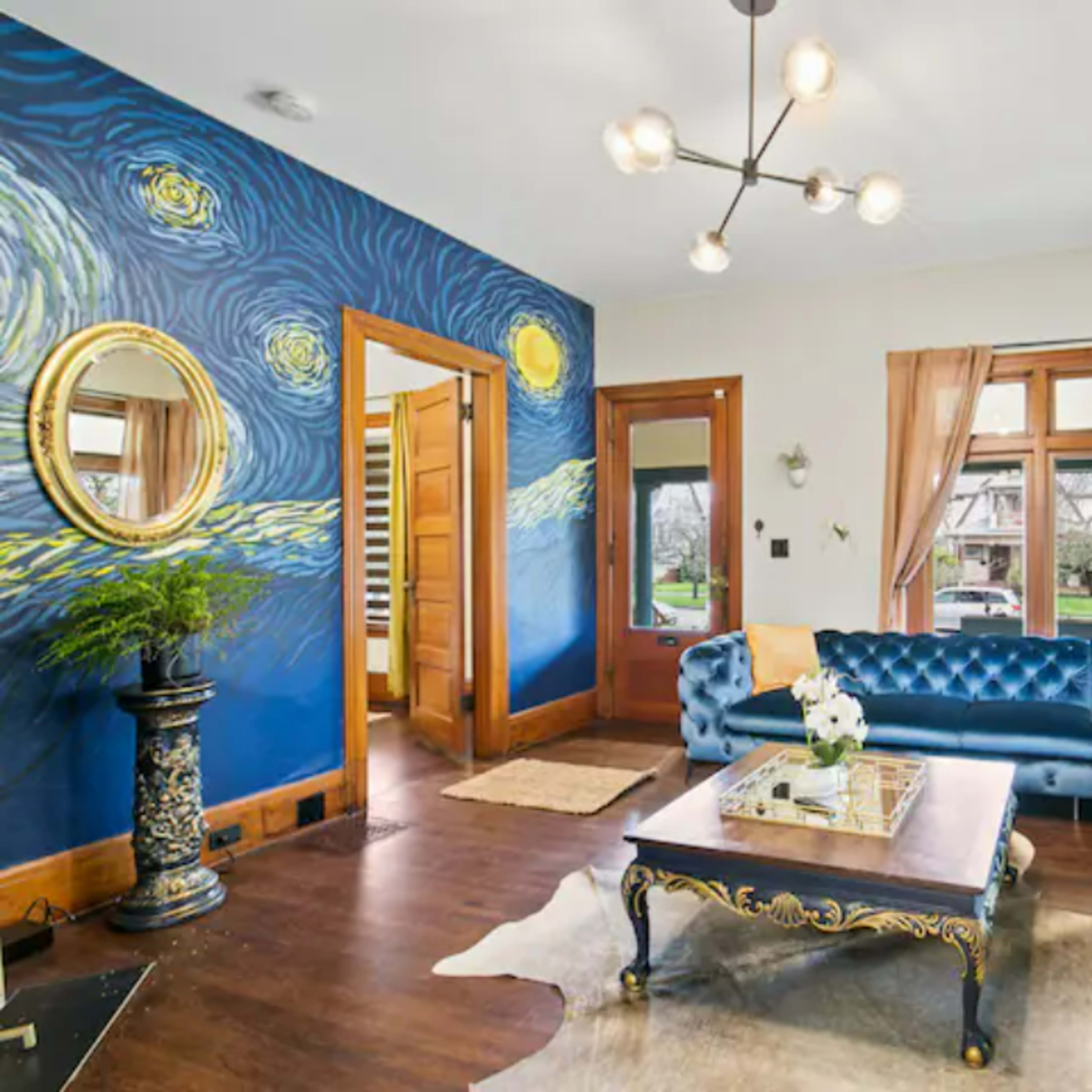 A living room features a blue velvet sectional sofa, a coffee table, and a mural of swirling stars on one wall, with large windows allowing natural light.