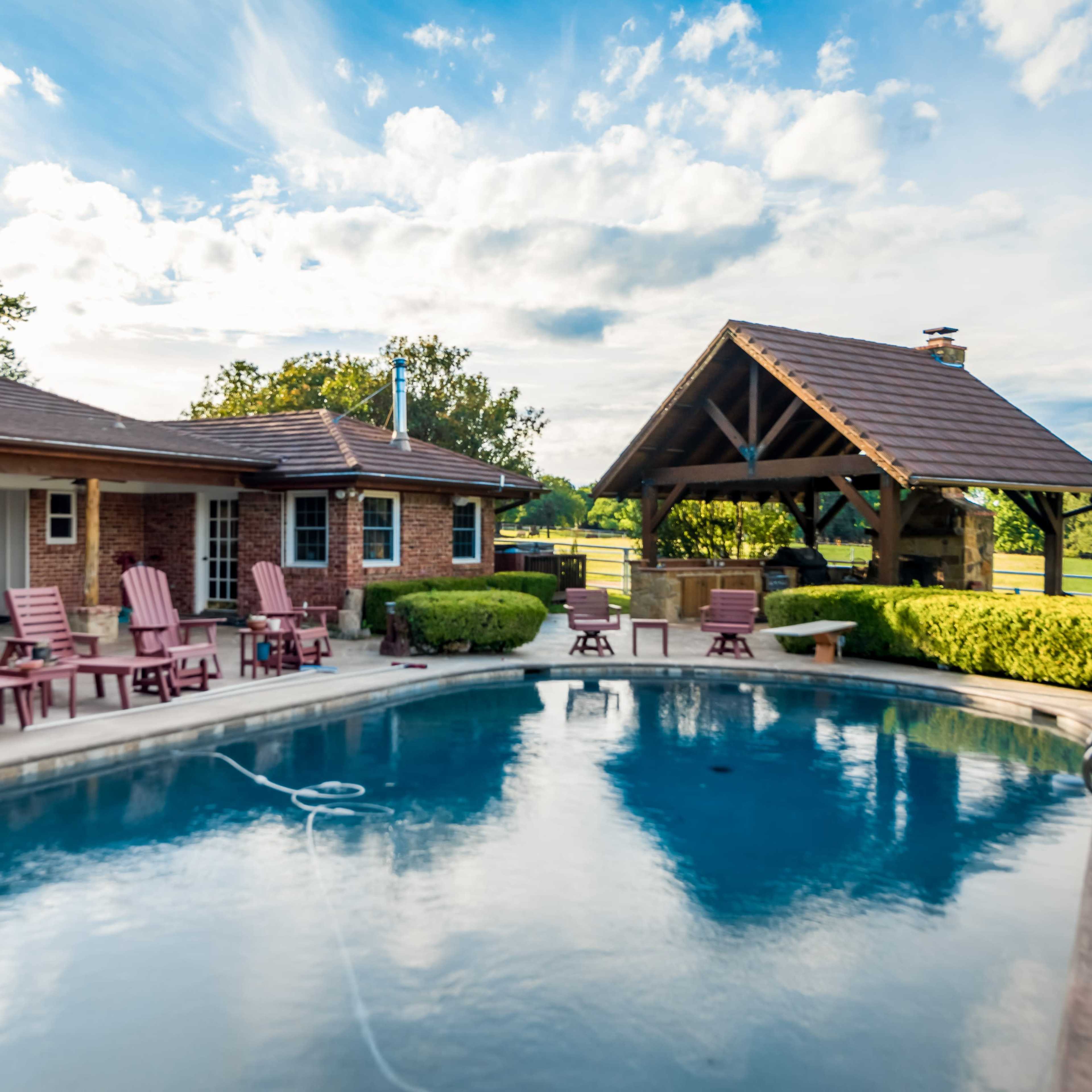 A backyard features a swimming pool surrounded by wooden lounge chairs and a pavilion with a sloped roof.