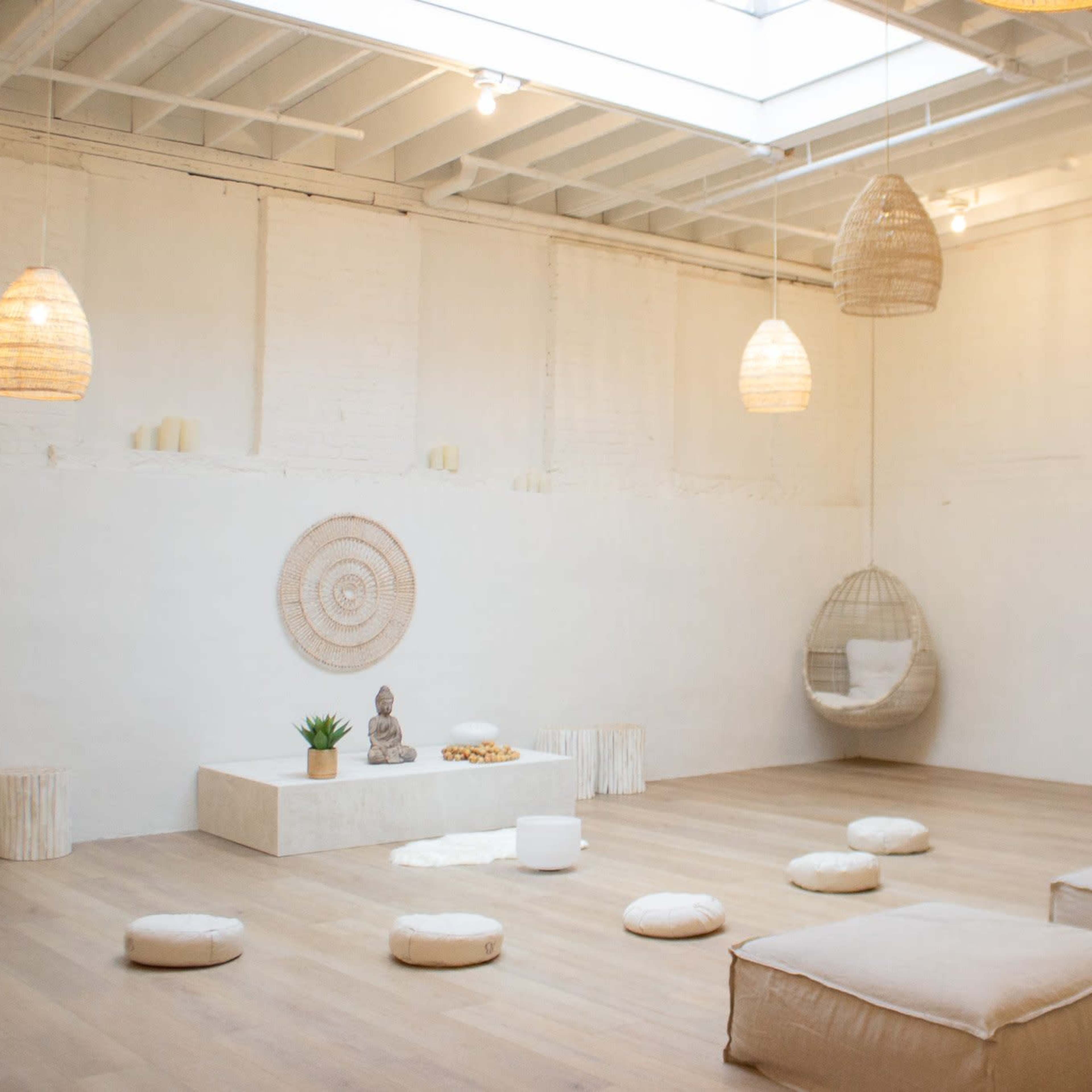 A minimalist meditation space with soft seating, a decorative wall hanging, a small statue, and plants, illuminated by natural light from a skylight.